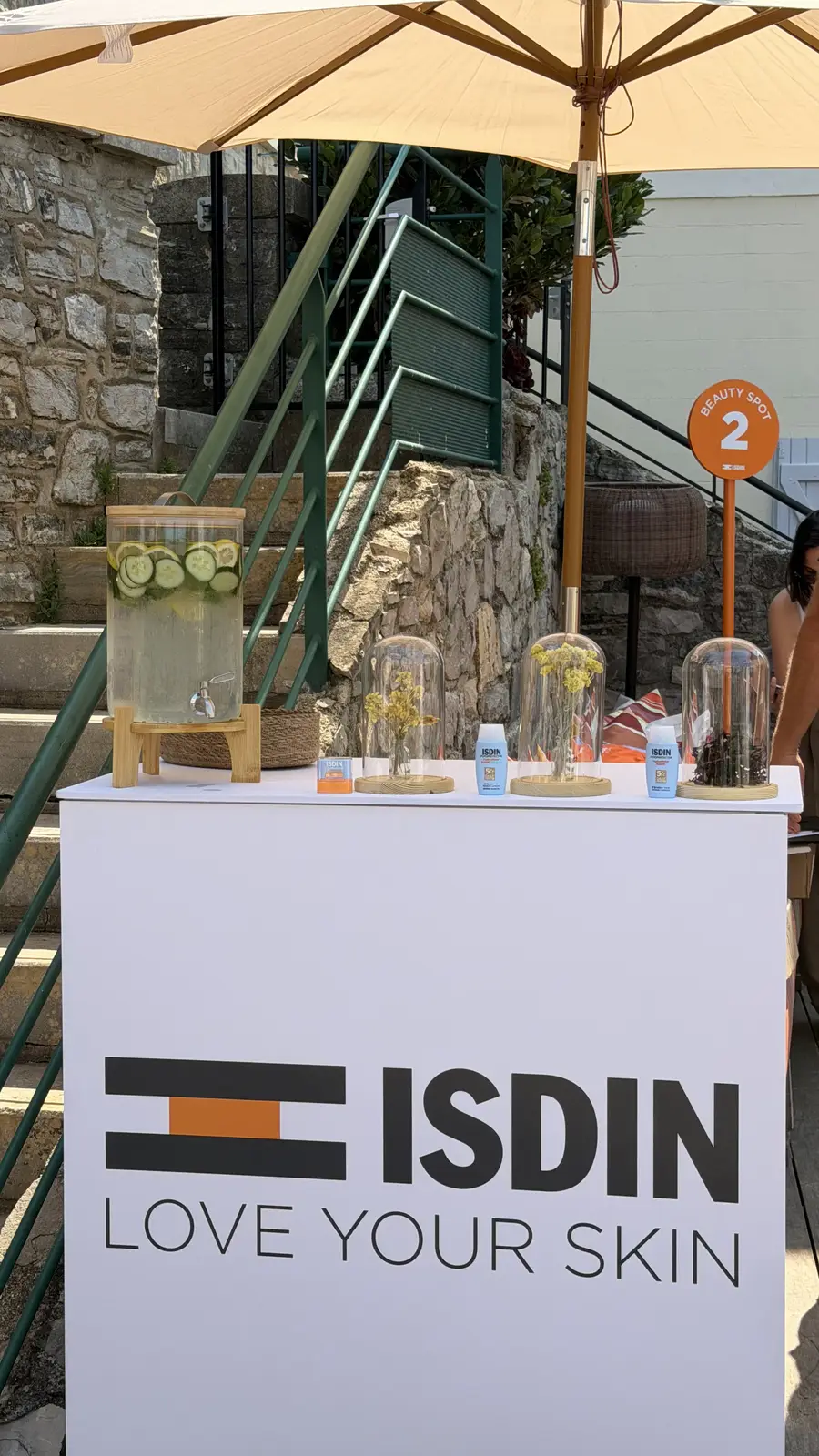 Présentoir blanc en extérieur avec le logo ISDIN et la phrase "Love Your Skin", contenant des produits cosmétiques sous cloches en verre et une grande carafe d'eau infusée avec des tranches de concombre et citron sous un parasol beige.