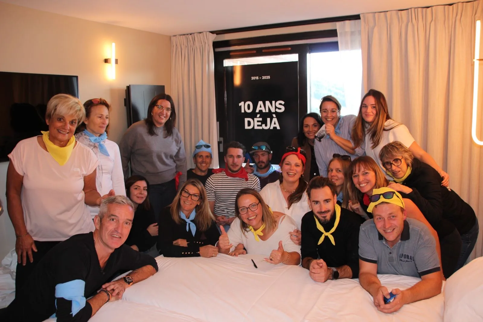 Groupe de 17 personnes souriantes, avec des foulards colorés autour du cou, posant dans une chambre d'hôtel devant une fenêtre avec une affiche indiquant « 10 ans déjà 2015 - 2025 ».