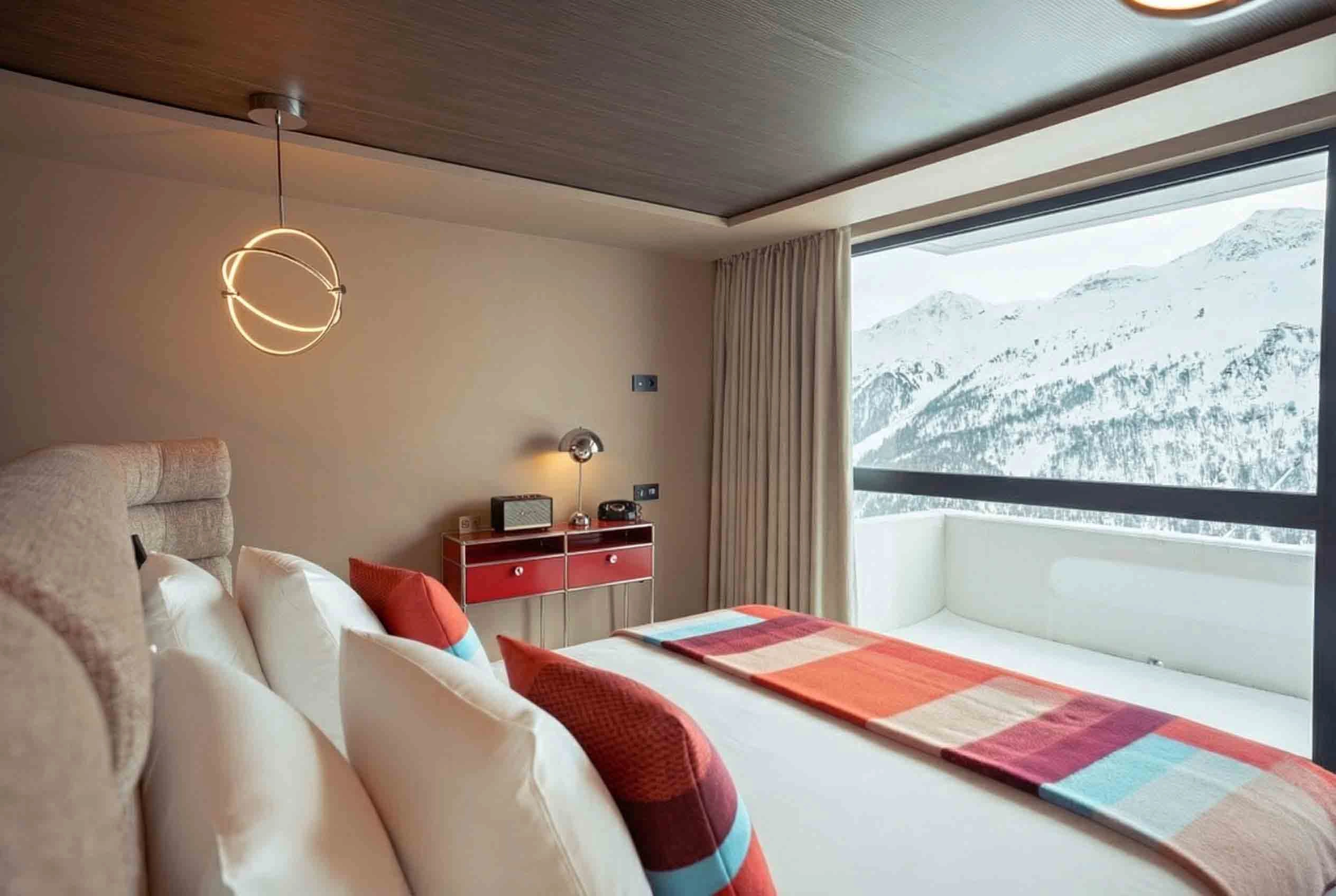 Chambre d'hôtel moderne avec un grand lit décoré de coussins et d'une couverture colorée, une table de chevet rouge avec une lampe et un téléphone, et une grande fenêtre offrant une vue sur des montagnes enneigées.