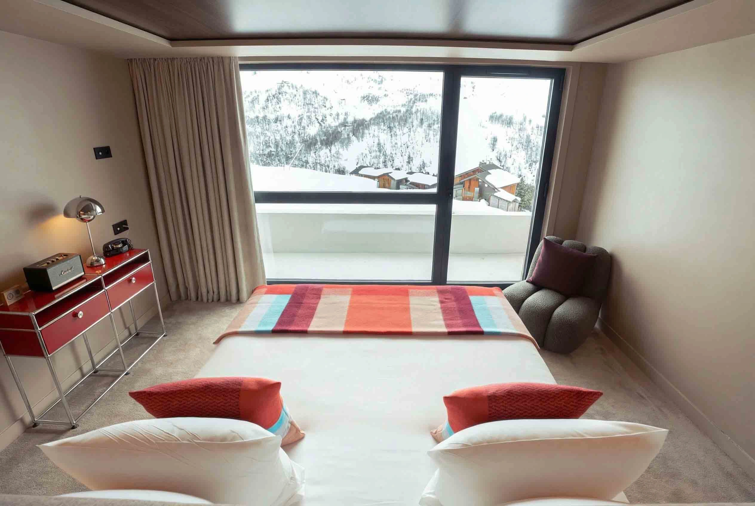 Chambre d'hôtel moderne avec un lit double blanc orné de coussins orange et bleus et d'un plaid rayé coloré, une chaise confortable grise dans un coin et une grande fenêtre donnant sur une vue enneigée de montagnes et de chalets en bois.