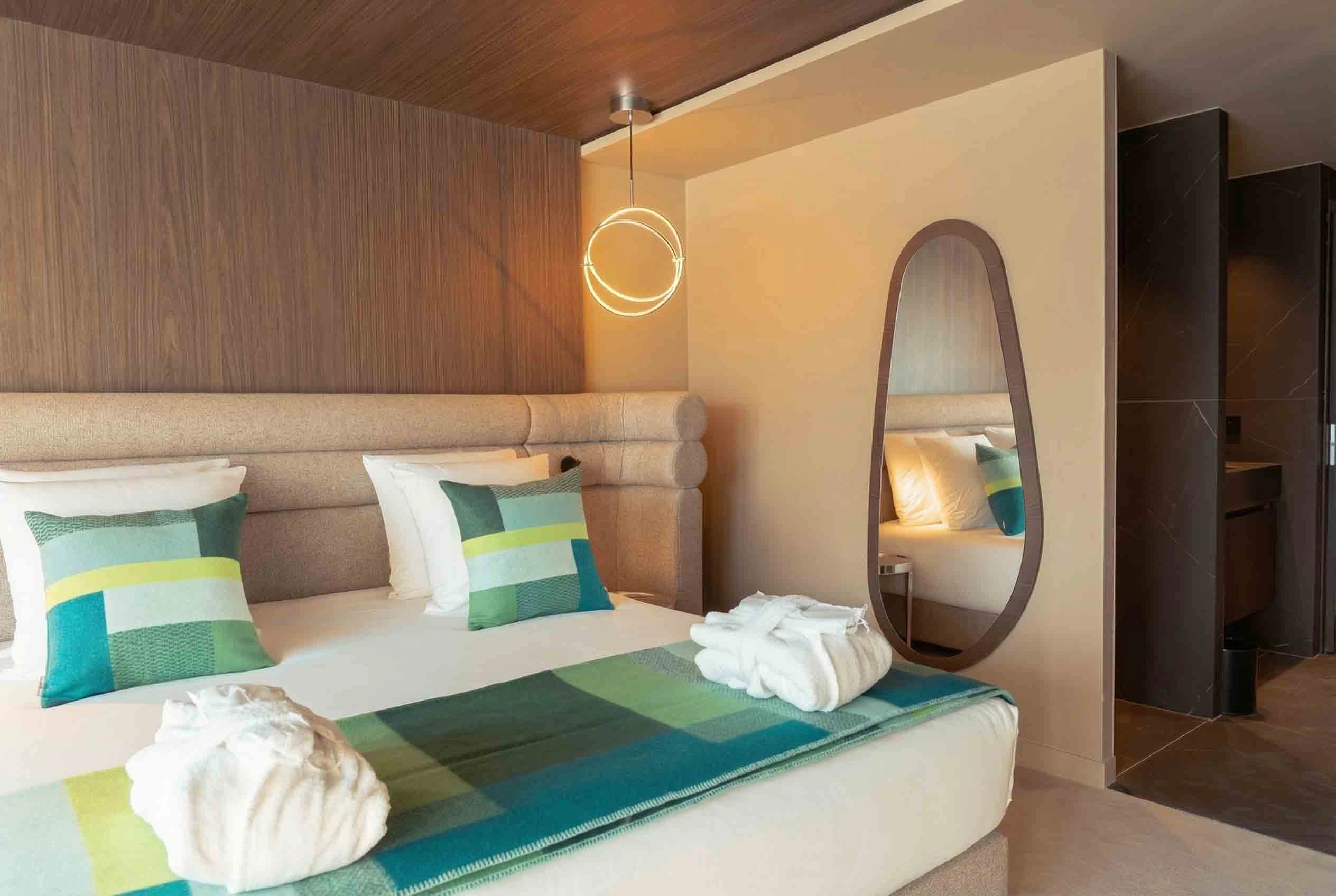 Chambre d'hôtel moderne avec un grand lit double, deux coussins aux motifs verts et bleus, deux peignoirs blancs pliés sur le lit, un mur en bois, un miroir mural ovale et un éclairage suspendu en forme d'anneau.