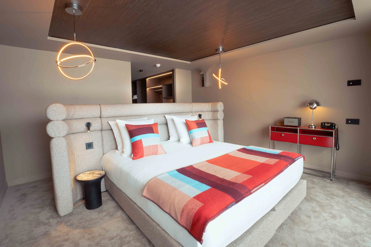 Chambre d'hôtel moderne avec un grand lit double beige, coussins colorés rouges, bleus et beiges, couverture géométrique rouge, orange, beige et bleu, table de chevet ronde noire et lampe suspendue circulaire.