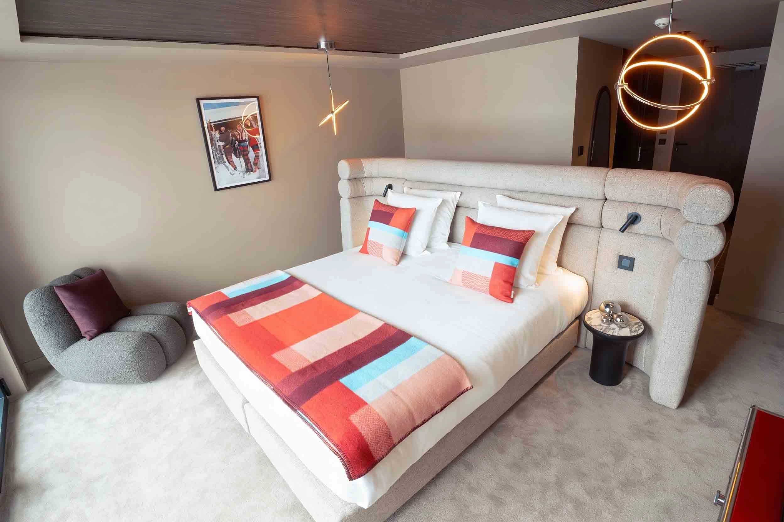 Chambre moderne avec un grand lit double orné de coussins rouges et d'une couverture multicolore, fauteuil gris avec coussin violet, table de chevet ronde avec décoration métallique, suspension lumineuse design au plafond et tableau mural illustrant des skieurs.