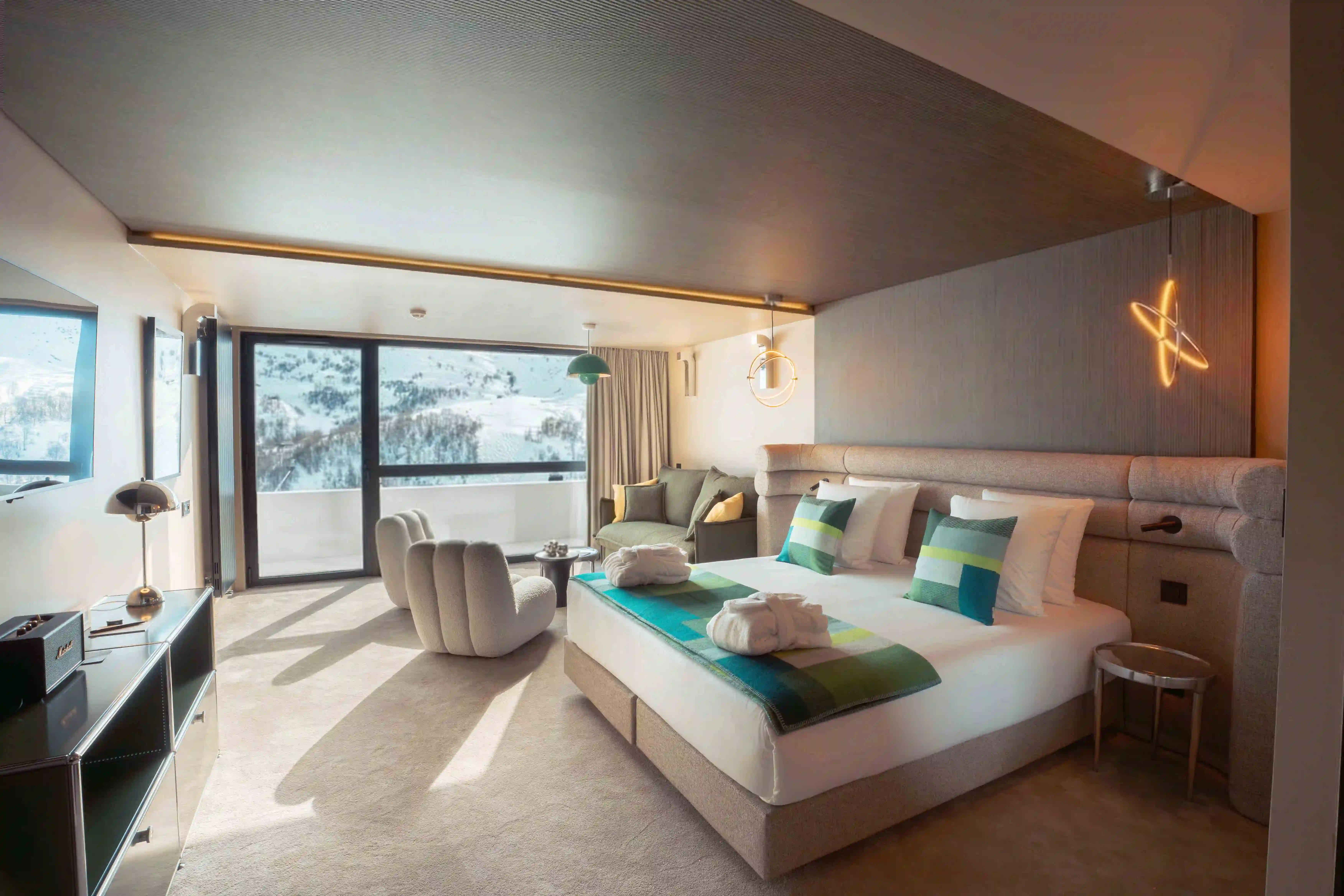Chambre d'hôtel moderne avec grand lit double décoré de coussins verts et blancs, deux peignoirs pliés, fauteuils beige en forme de nuage, canapé vert avec coussins, grande fenêtre panoramique montrant une vue sur une montagne enneigée, éclairage suspendu design, tapis beige et mobilier contemporain.