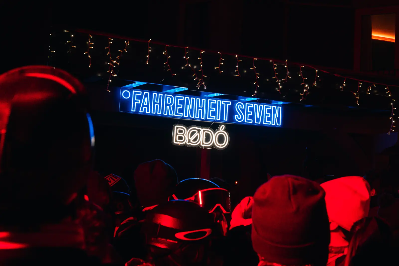 Une foule avec bonnets et casques dans une lumière rouge, sous un panneau lumineux bleu et blanc indiquant «FAHRENHEIT SEVEN BØDØ», décoré de guirlandes lumineuses.