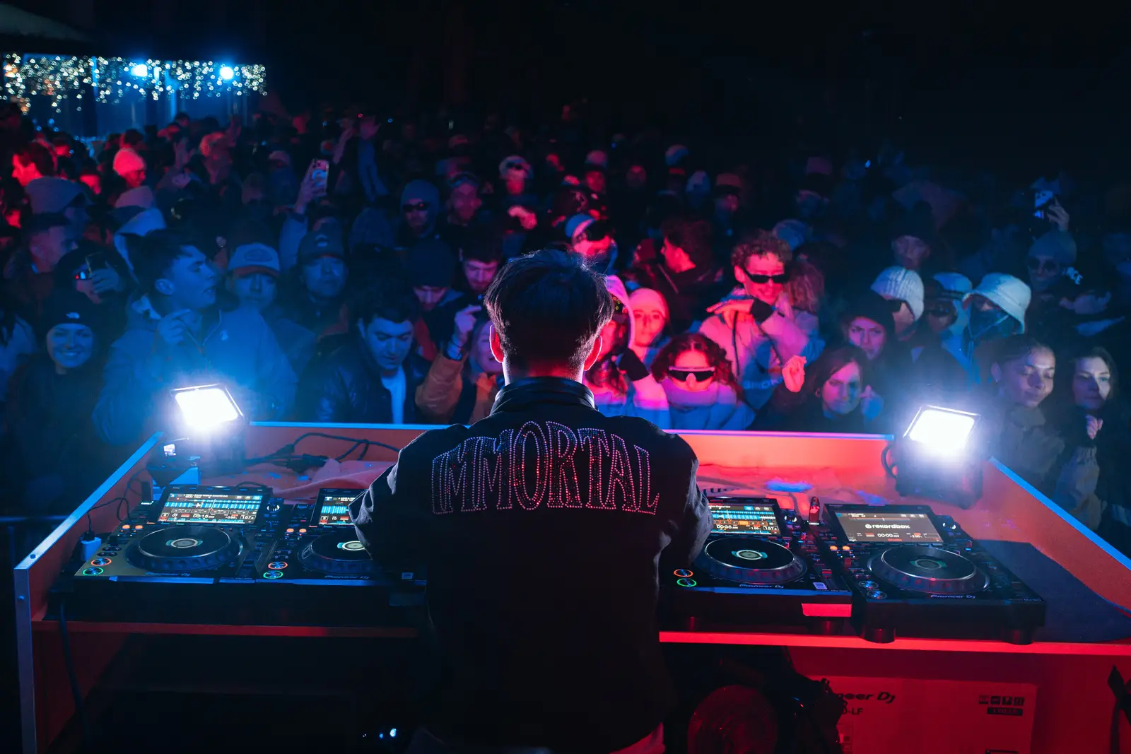 Un DJ vu de dos portant une veste avec le mot "IMMORTAL" en lettres lumineuses, joue de la musique sur des platines lors d'une fête nocturne en plein air, devant une foule attentive et éclairée en bleu et rouge.