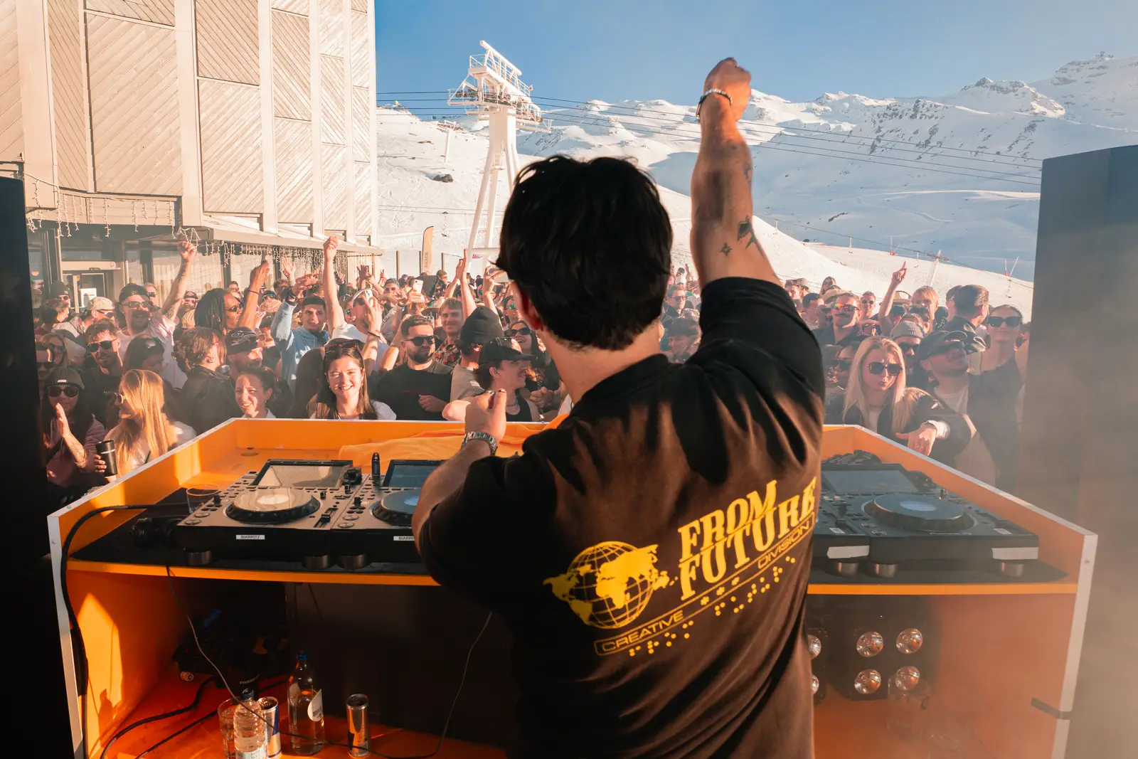 Un DJ vu de dos joue de la musique devant une foule enthousiaste à l'extérieur dans un paysage enneigé de montagnes, avec un ciel bleu clair.
