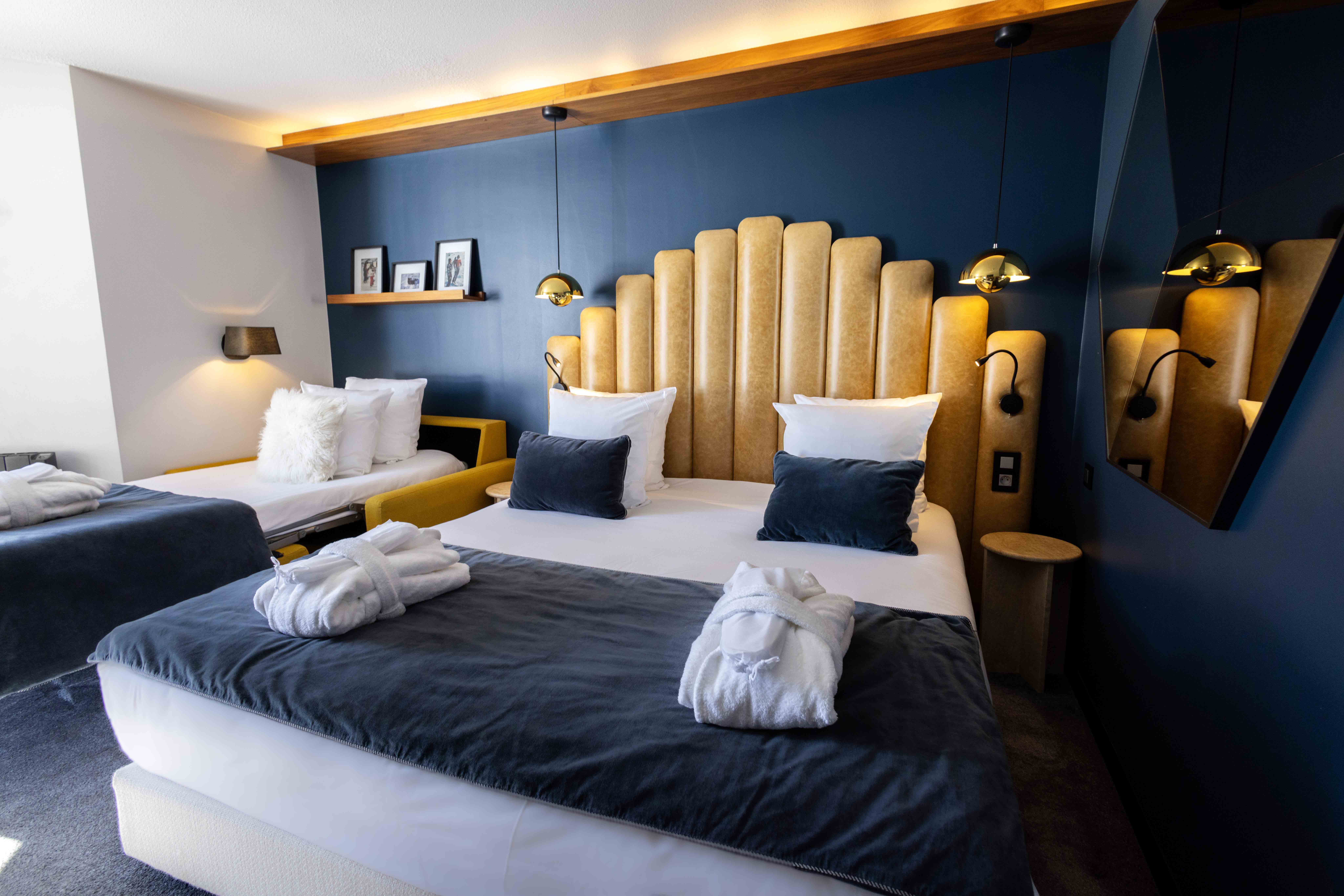 Chambre moderne avec un grand lit double muni d'une tête de lit beige matelassée, deux coussins bleus, et une couverture grise avec peignoirs blancs pliés dessus; un canapé-lit jaune avec coussins blancs à gauche, murs bleu foncé avec lampe murale et deux suspensions dorées, et un miroir incliné à droite.