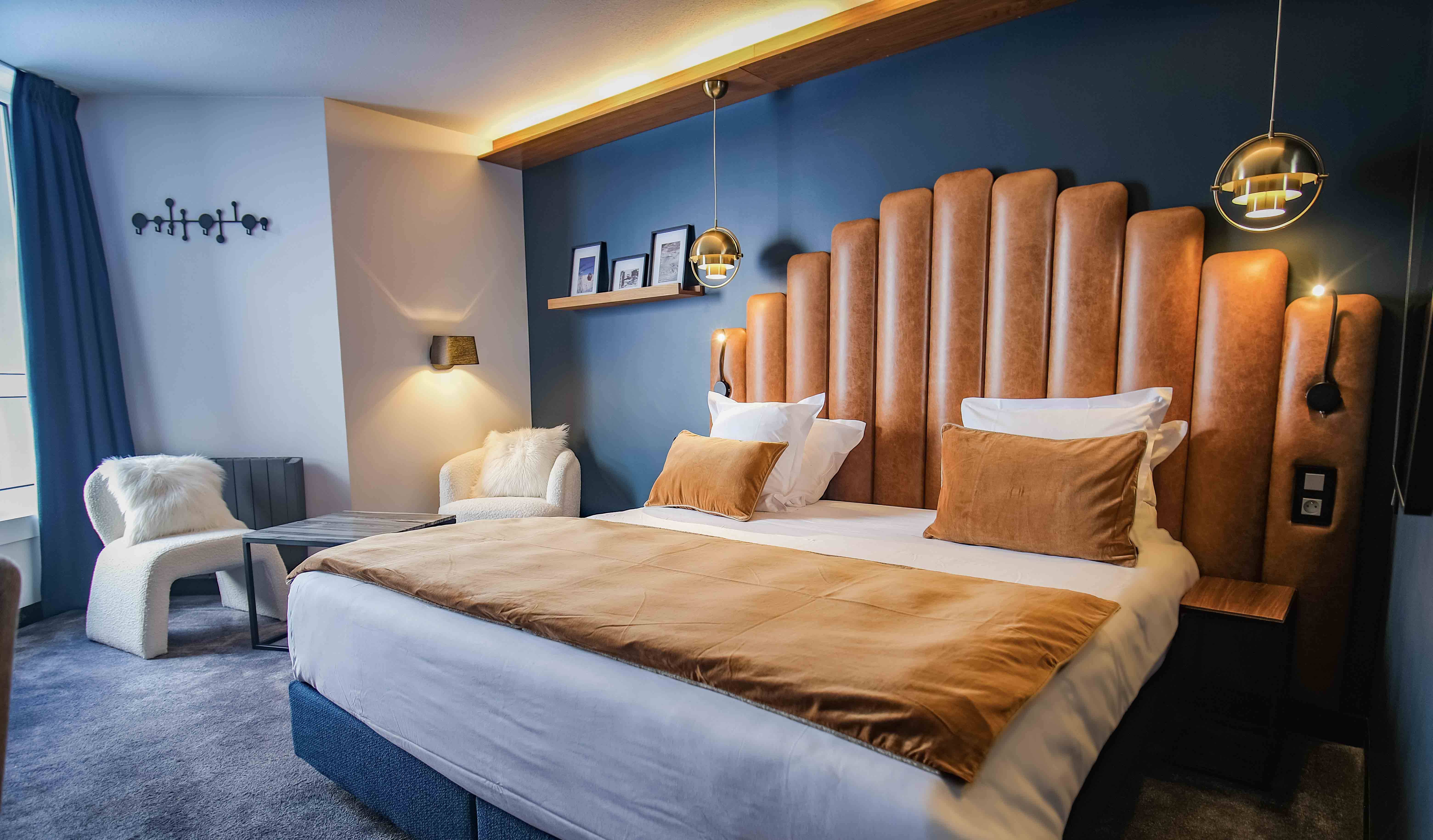 Chambre d'hôtel moderne avec un grand lit double doté d'une tête de lit en cuir marron côtelée, deux coussins et une couverture beige, deux fauteuils blancs moelleux avec coussins, une petite table basse, des rideaux bleus, un mur bleu foncé avec une étagère portant trois cadres photo et deux suspensions lumineuses rondes en métal doré.