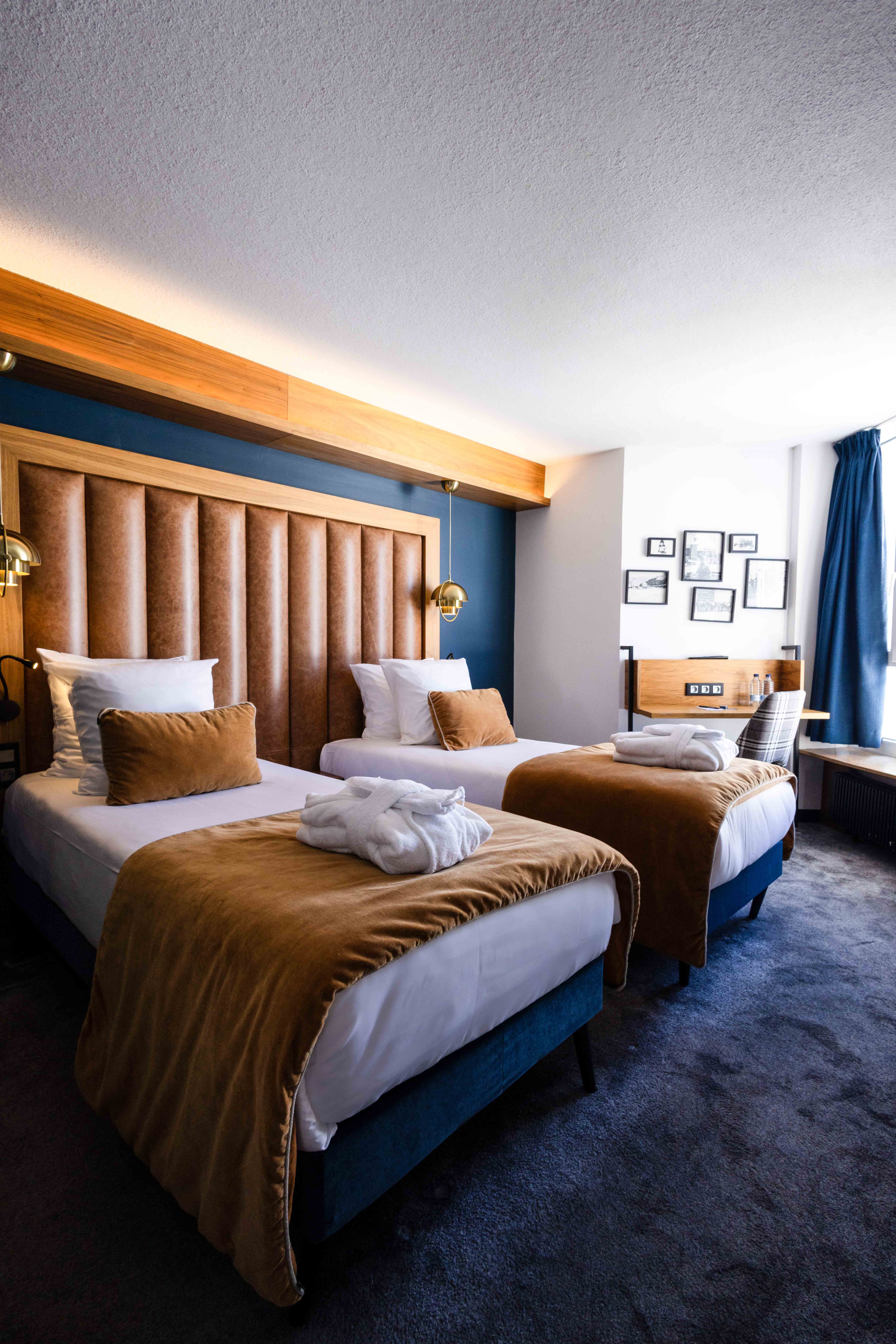 Chambre d'hôtel avec deux lits simples coiffés de couvertures marron, coussins assortis, têtes de lit rembourrées en cuir marron, peignoirs blancs pliés sur chaque lit, moquette sombre, bureau en bois avec chaise à motifs, et rideaux bleus près d'une fenêtre.