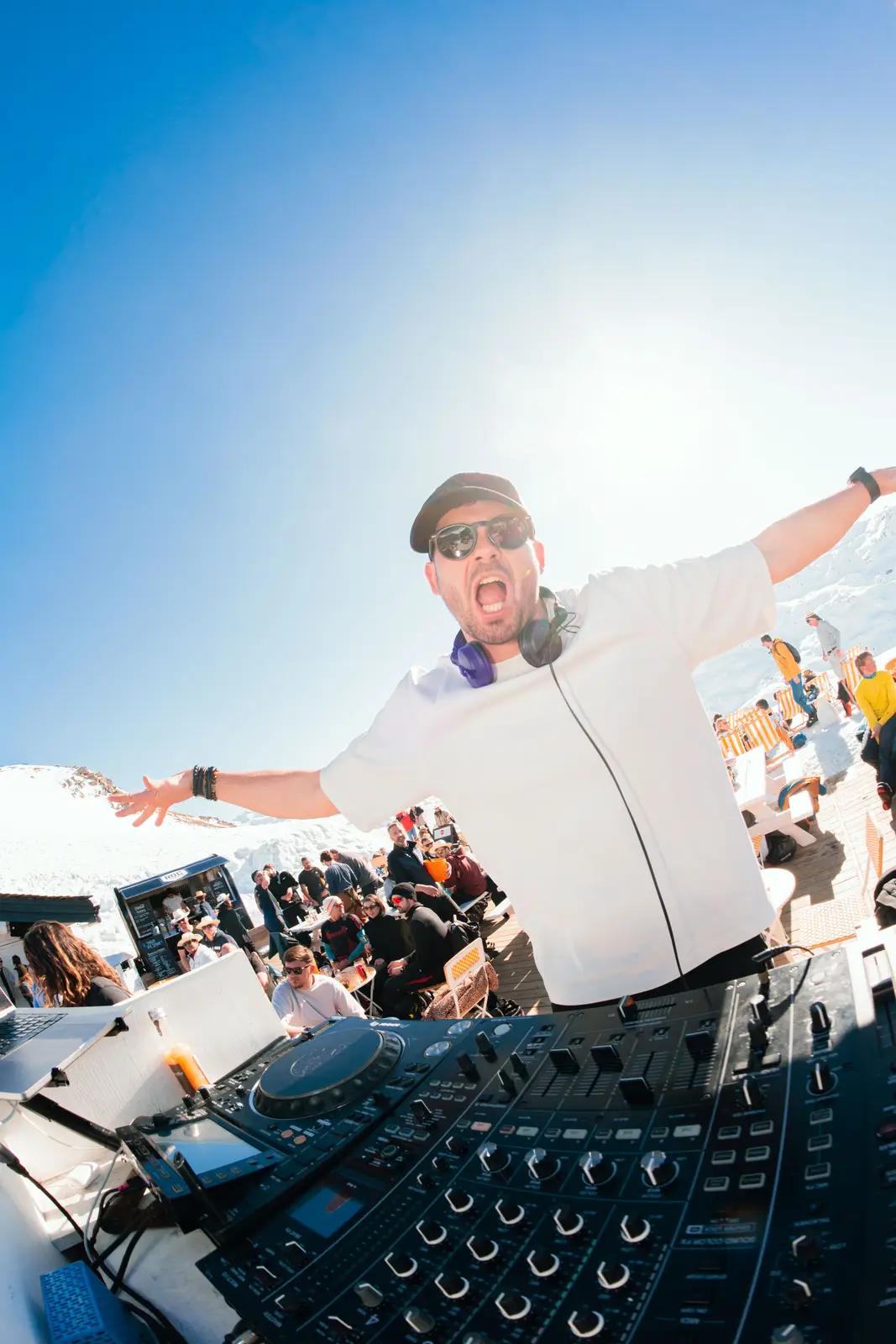 Un DJ souriant portant une casquette, des lunettes de soleil et un casque autour du cou, les bras écartés, devant une console de mixage en plein air sous un ciel bleu, avec un public assis et debout sur une terrasse enneigée.