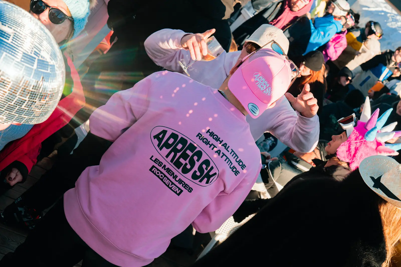 Person wearing a pink sweatshirt and matching cap with text 'APRES SKI LES MENUIRES ROC SEVEN' and 'HIGH ALTITUDE RIGHT ATTITUDE' at a lively outdoor winter party with people dressed in colorful winter clothes, including one person with a unicorn hat and another holding a disco ball.