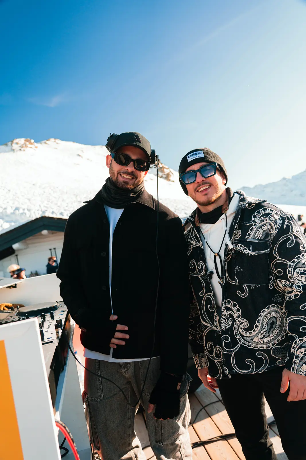 Deux hommes souriants portant des lunettes de soleil et des vêtements d'hiver posent côte à côte sur une terrasse en bois avec de la neige et des montagnes en arrière-plan par une journée ensoleillée.