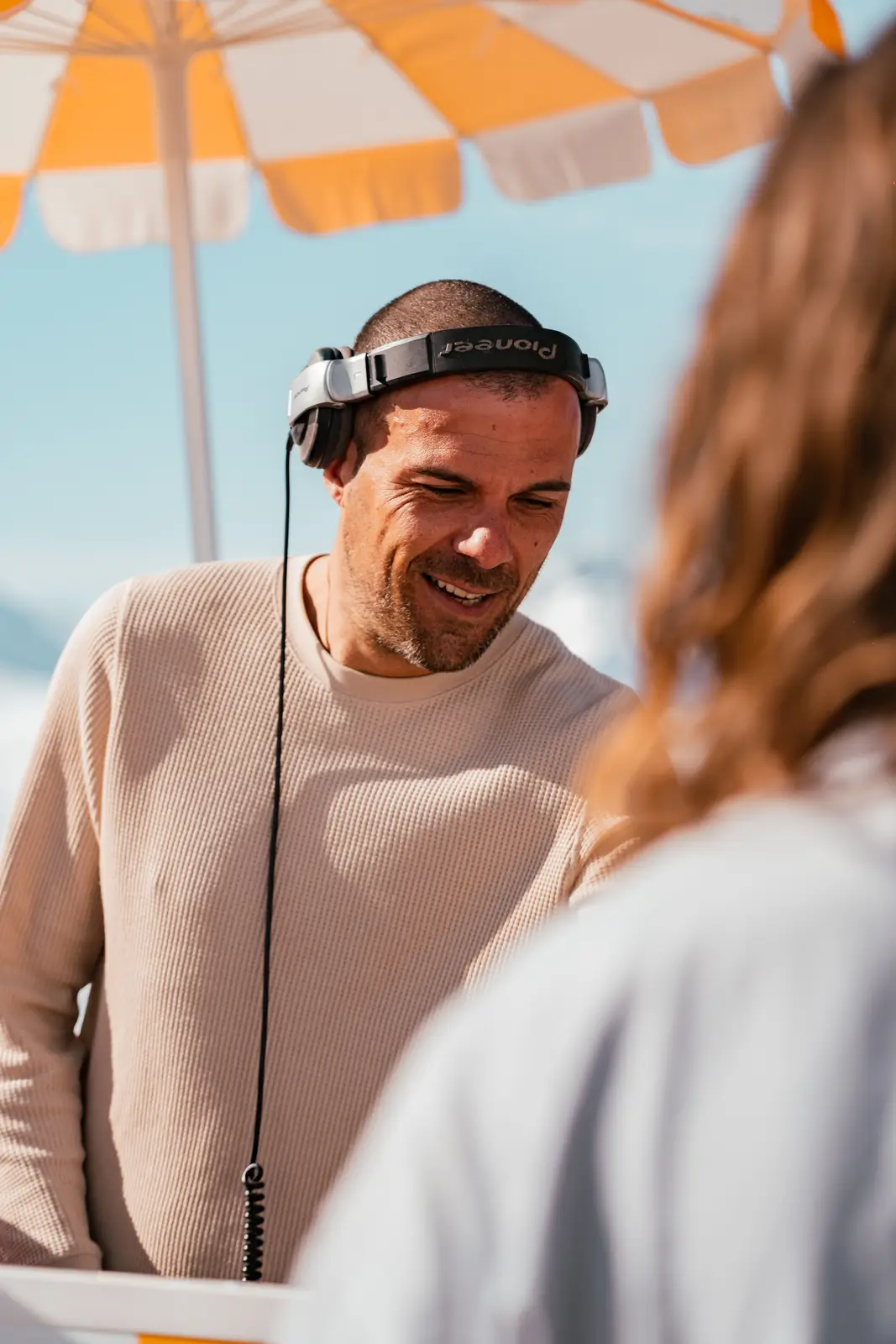 Homme portant un pull beige et un casque audio Pioneer, debout sous un parasol orange et blanc lors d'une journée ensoleillée, parlant avec une autre personne floue au premier plan.