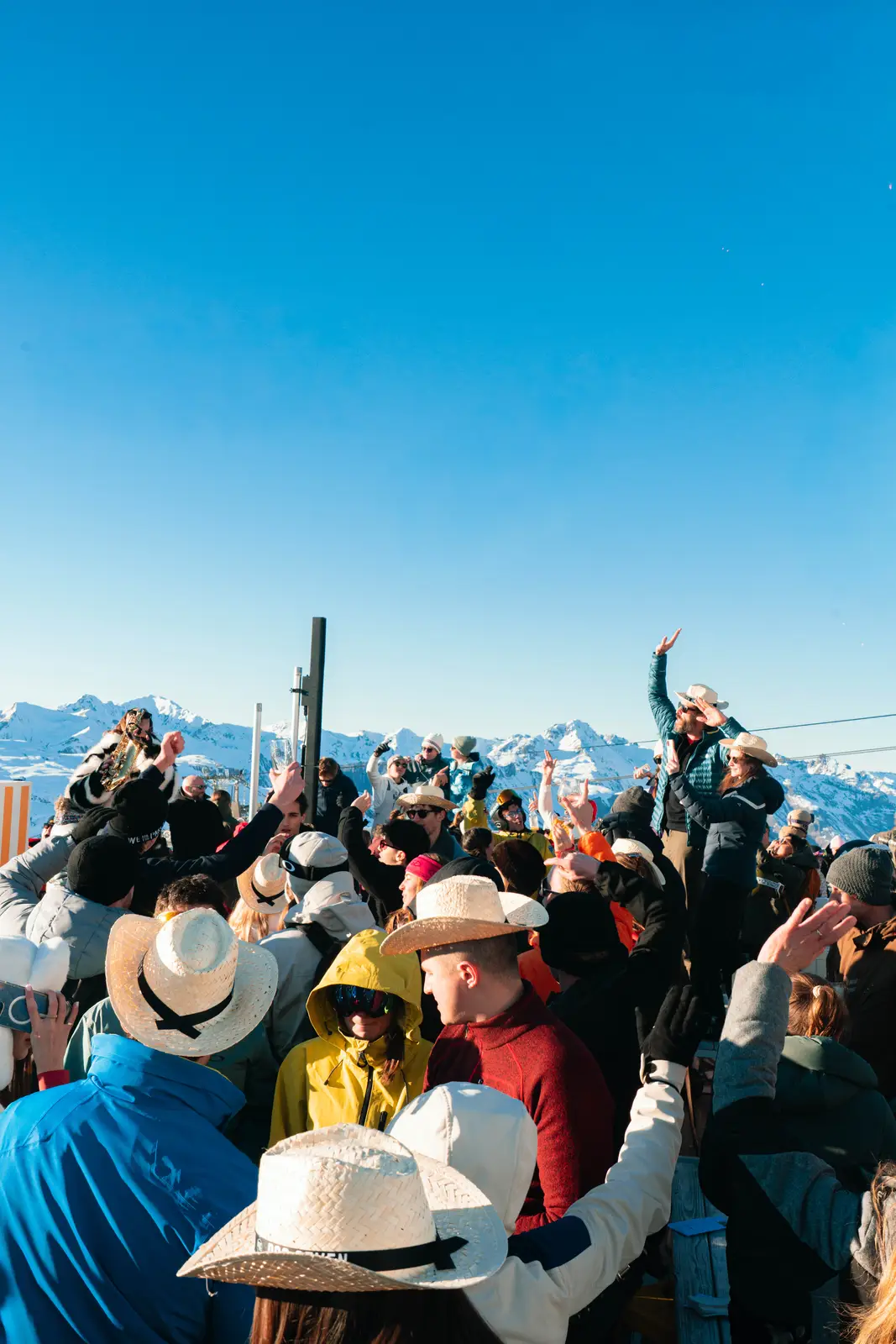 Groupe de personnes en tenue d'hiver avec des chapeaux de paille, certaines levant les bras, faisant la fête à l'extérieur avec des montagnes enneigées en arrière-plan sous un ciel bleu clair.