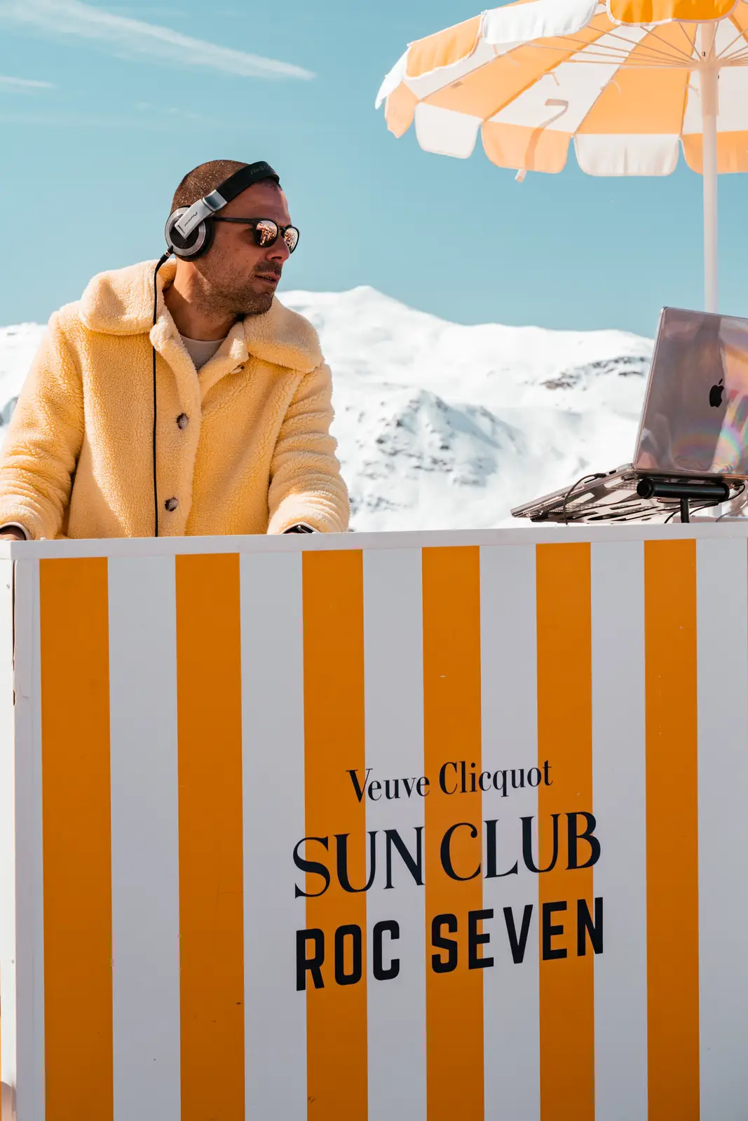 Un homme portant un manteau jaune et des lunettes avec des écouteurs anime en plein air derrière un stand à rayures blanches et orange portant le texte Veuve Clicquot Sun Club Roc Seven avec montagnes enneigées en arrière-plan et parapluie orange et blanc.