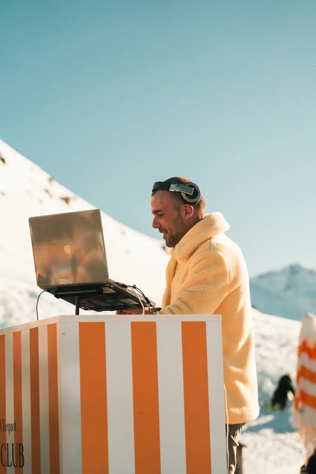 Un homme portant un casque audio et une veste en laine jaune regarde un ordinateur portable sur une station de DJ rayée orange et blanche avec un paysage de montagne enneigée en arrière-plan sous un ciel bleu clair.