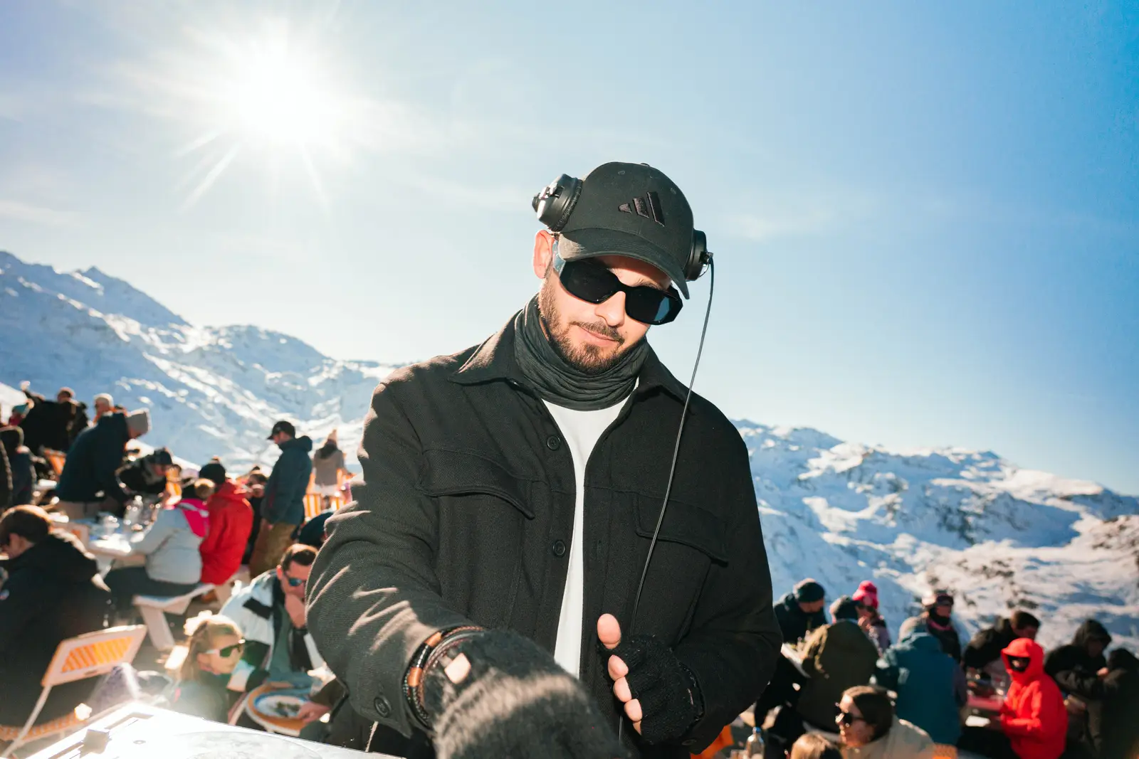 Un DJ portant une casquette noire, des lunettes de soleil et des gants, joue de la musique en extérieur dans une station de ski ensoleillée avec des montagnes enneigées et un groupe de personnes en arrière-plan.