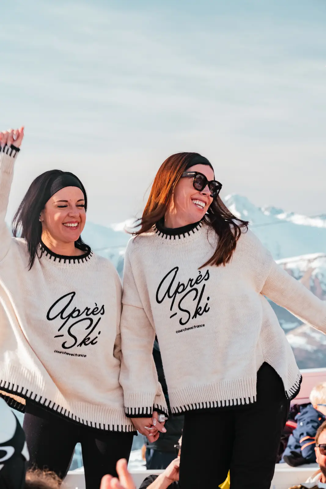 Deux femmes portant des pulls assortis crème avec l'inscription "Après-Ski Courchevel France", tenant la main et souriant devant un paysage de montagnes enneigées.