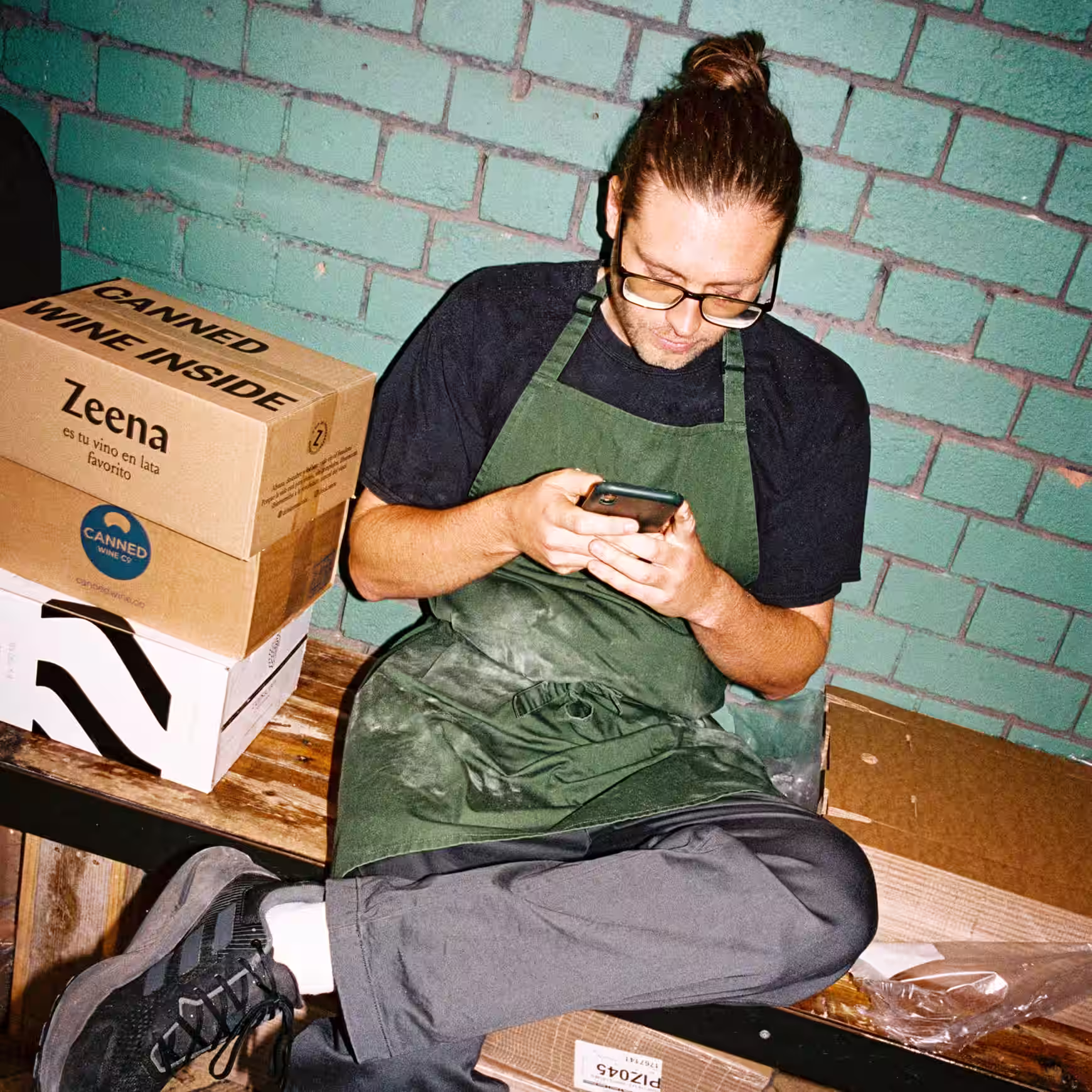 Restaurant staff member in green apron taking a break and using a phone in a storage area with boxed supplies