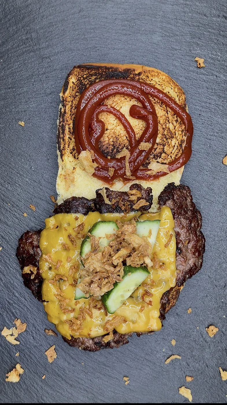 Aufgeklappter Smashed Burger von oben: getoastetes Bun mit Ketchup, Patty mit Cheddar, Röstzwiebeln und Gurken