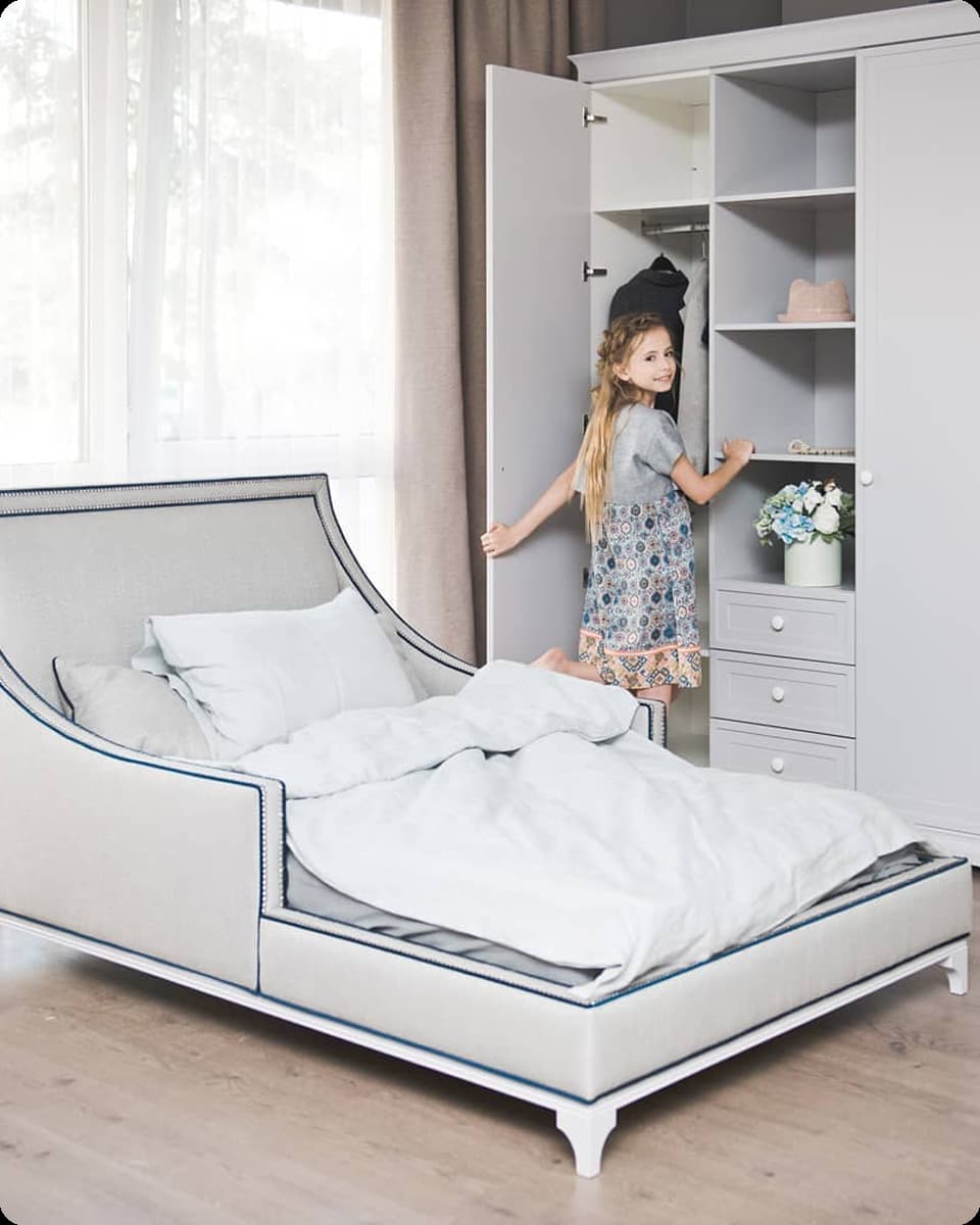 Little girl opening white wardrobe door in a bright bedroom with unmade grey bed.