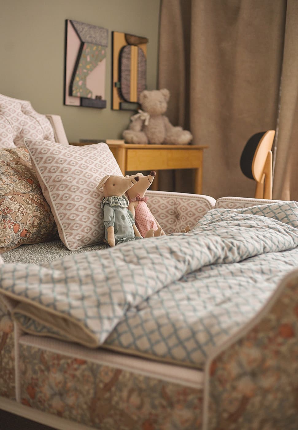 Patterned bed with two stuffed mice toys sitting on pillows and a teddy bear on a wooden desk behind it.