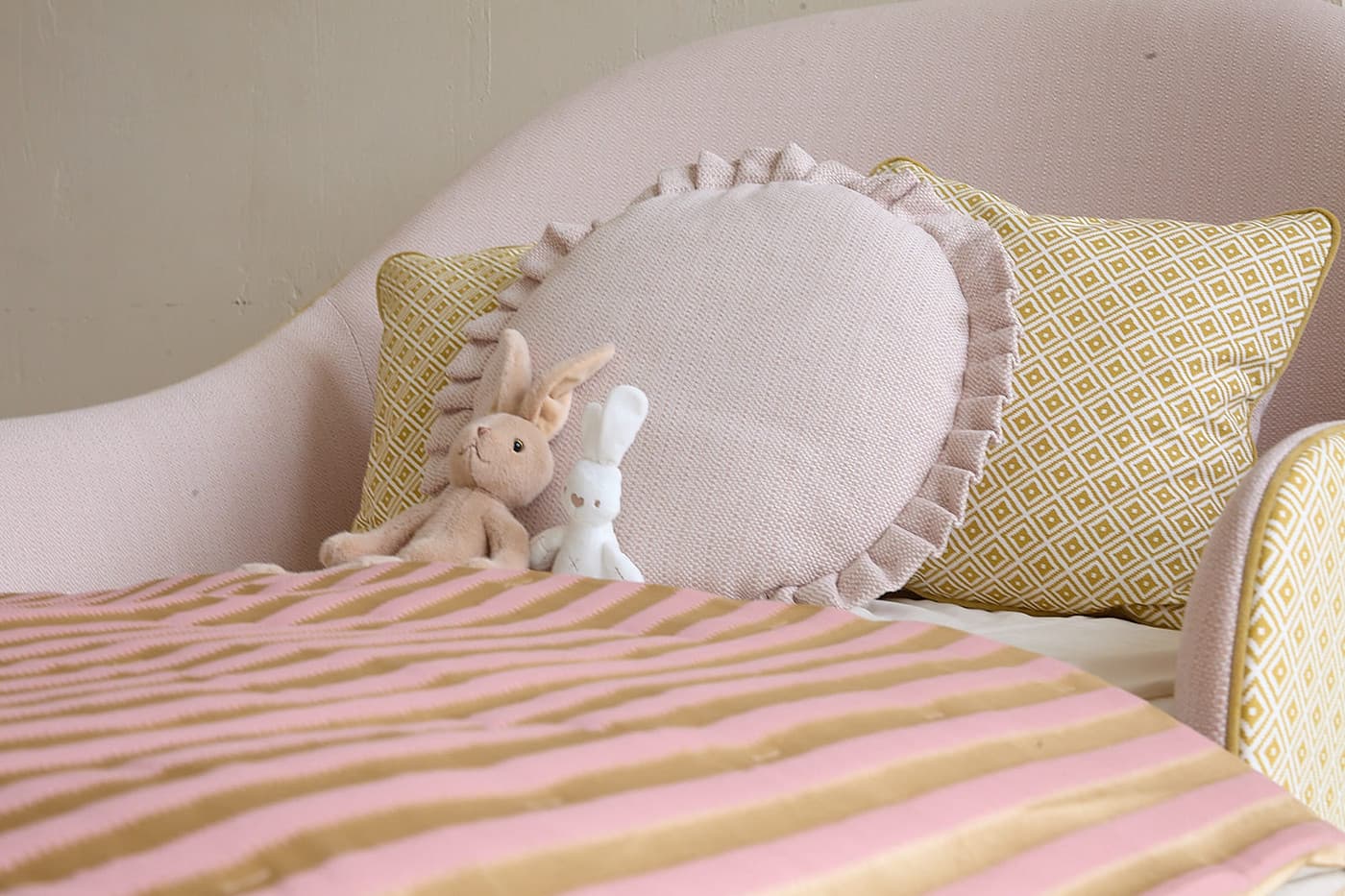 Pink and beige bed with two stuffed bunny toys and patterned pillows on a soft headboard.