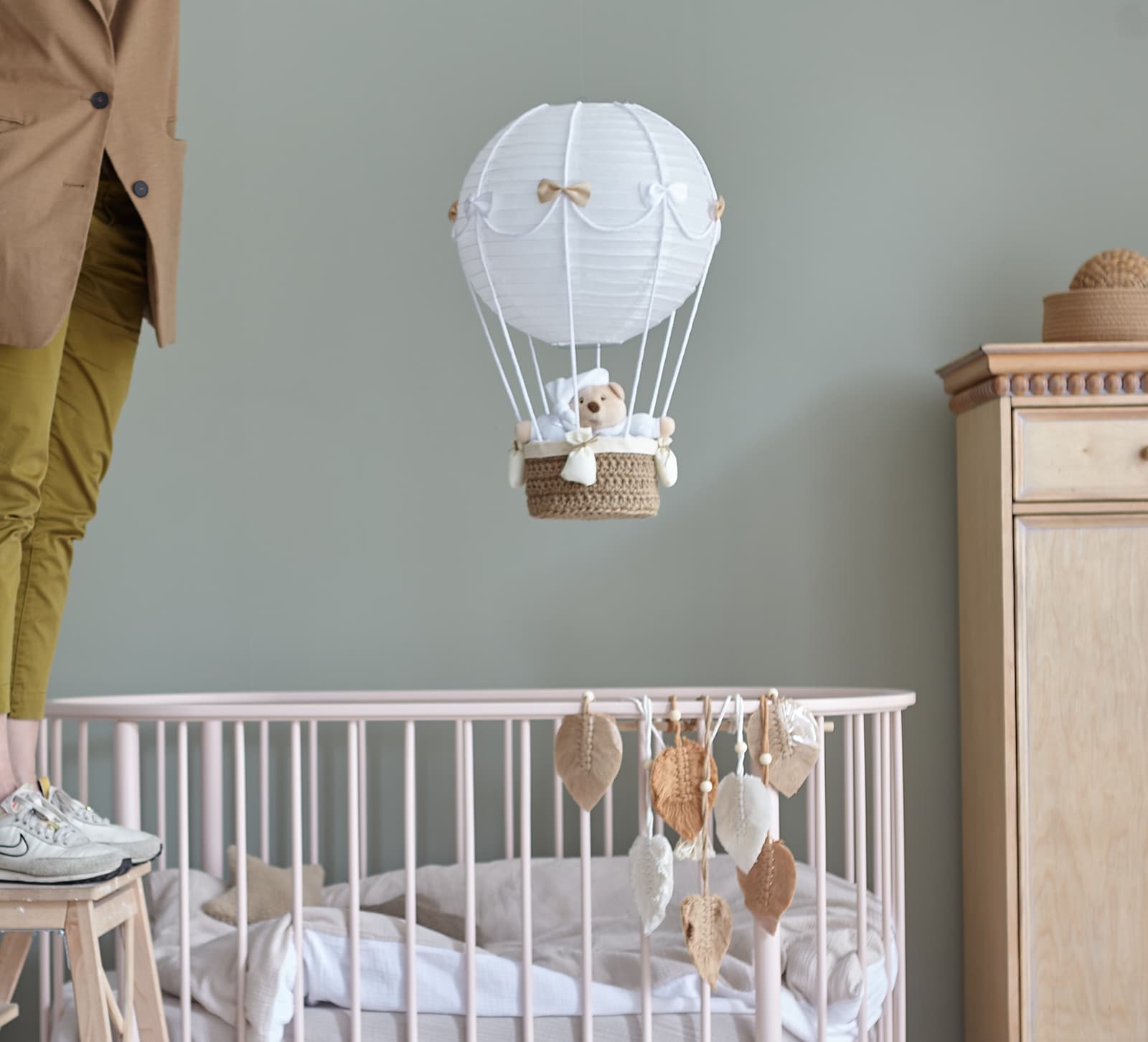 Baby crib with hanging leaf mobile and a teddy bear in a hot air balloon decoration above it.