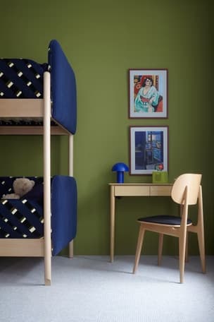 Room with wooden bunk bed, teddy bear, olive wall, desk with blue lamp, chair, and two framed artworks.