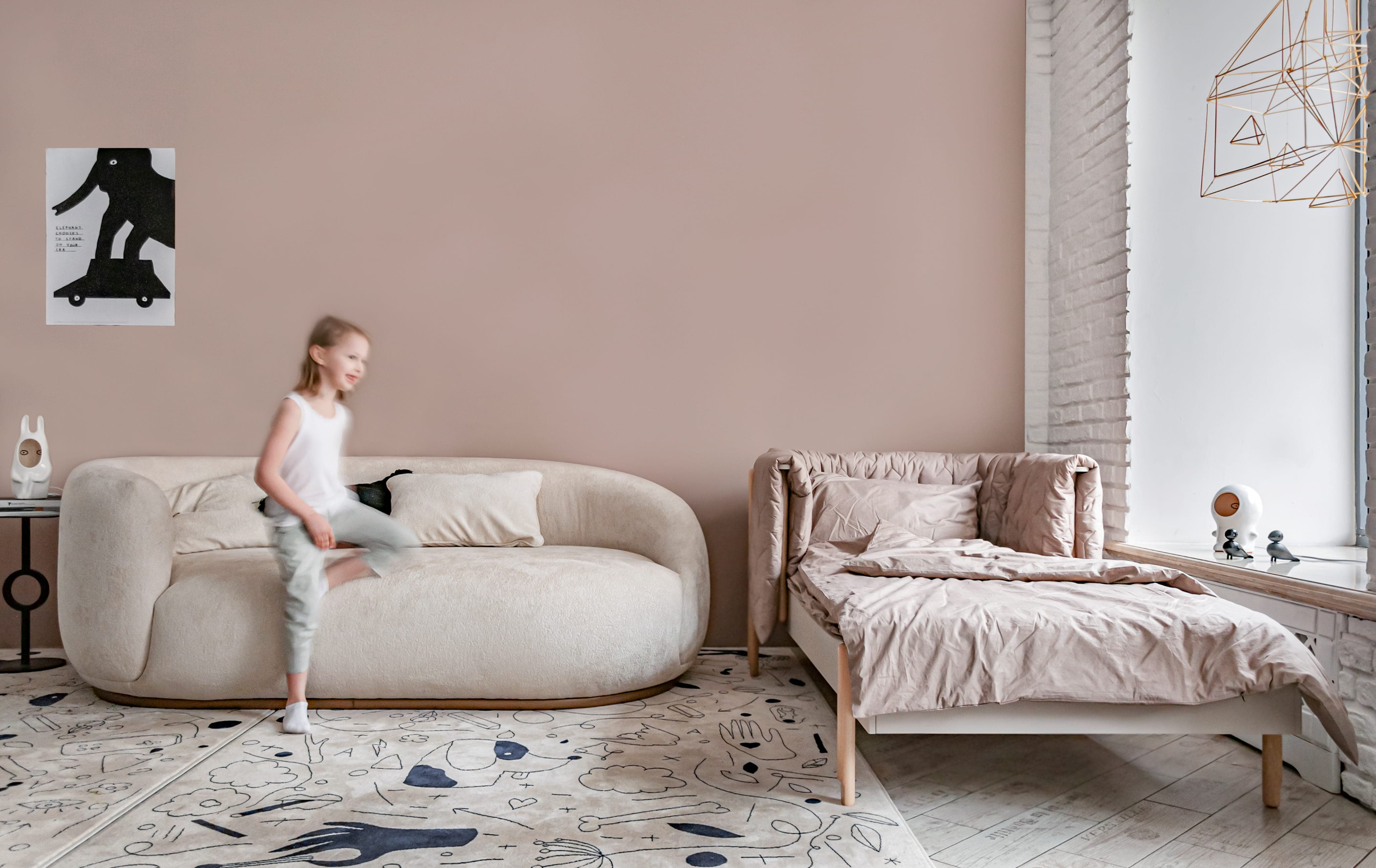 Child in white sleeveless shirt and pants walking in a cozy room with beige sofa and bed.
