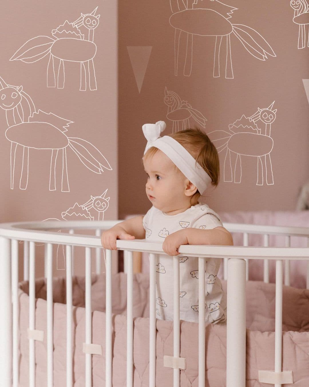 Baby wearing white headband standing and holding the rail of a white crib with unicorn wall art.