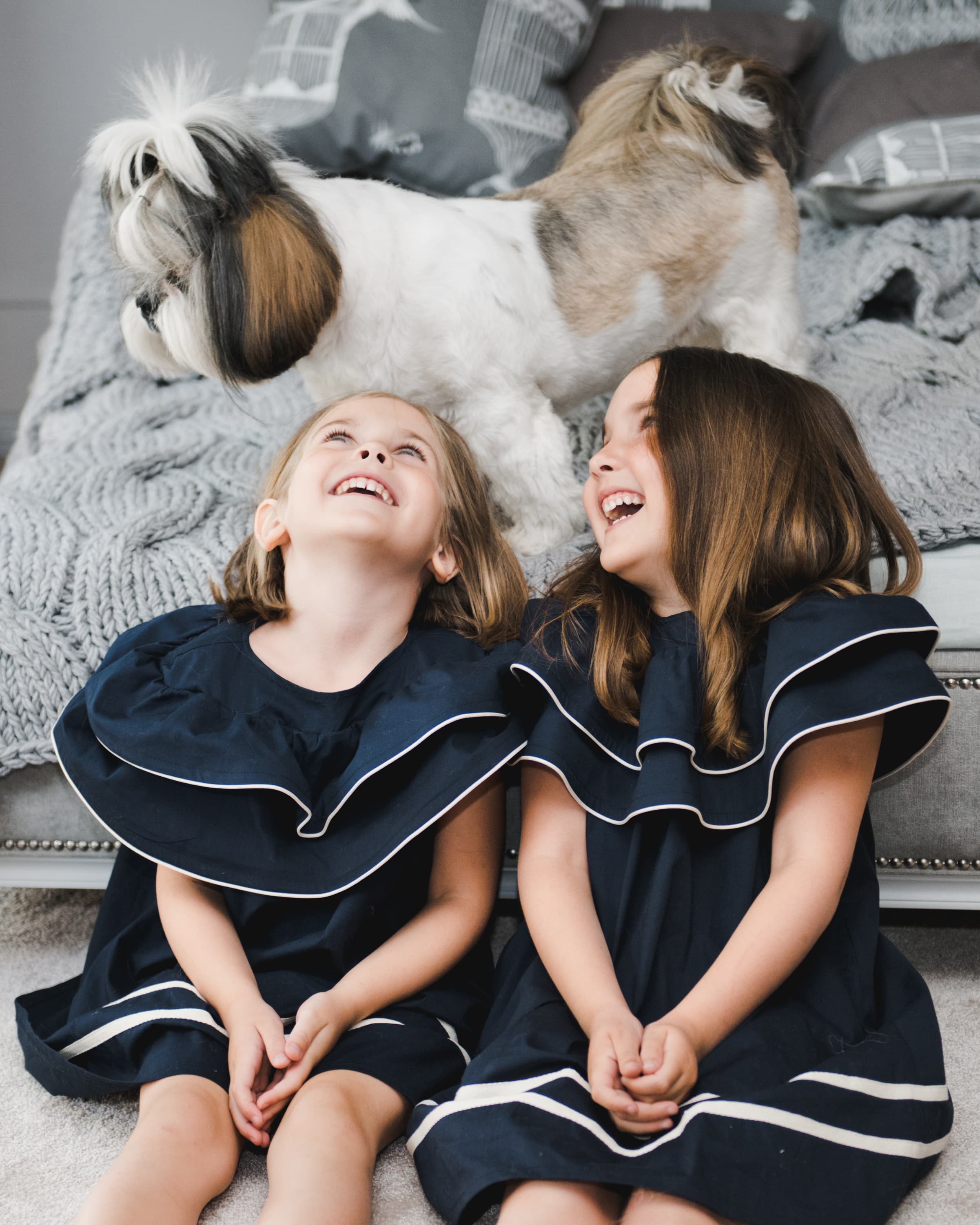 Two girls in matching navy dresses sitting on the floor laughing, with a small dog on the couch behind them.