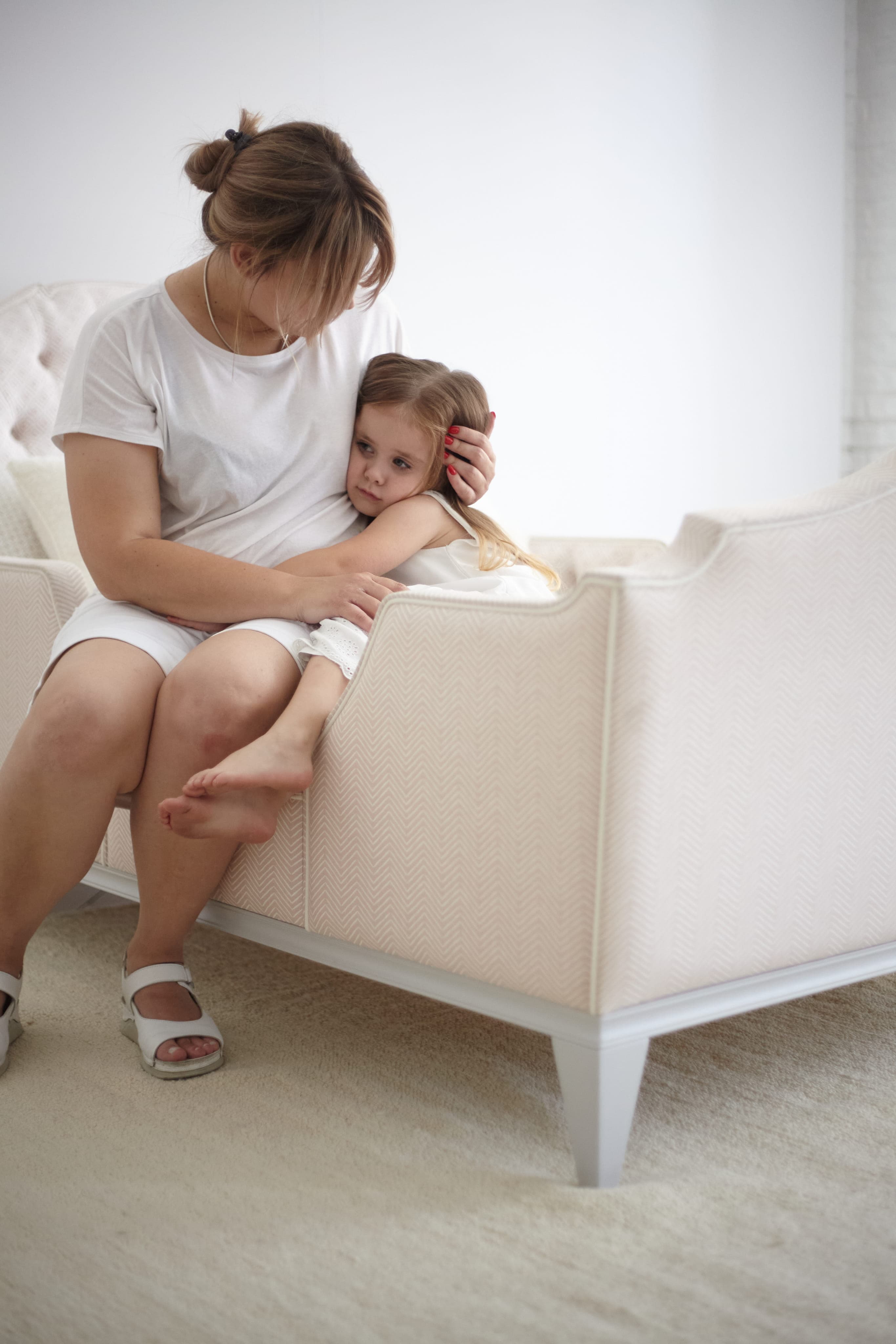 Woman in white clothes comforting a sad little girl sitting on a pale pink couch in a bright room.