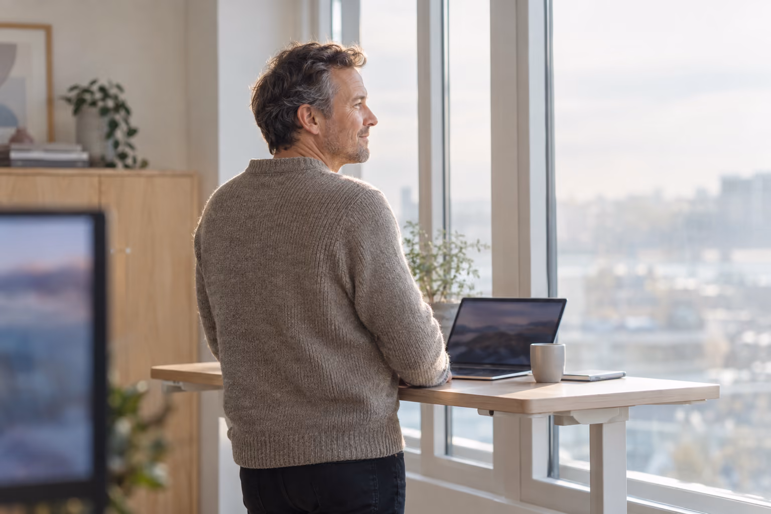 Ein Mann steht in hellem Büro vor großer Fensterfront und schaut nachdenklich aus dem Fenster. Vor ihm ein Stading-Desk mit einen Laptop.
