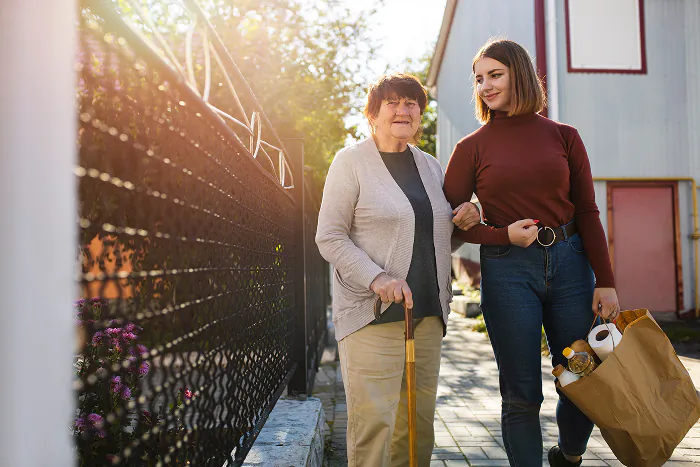 Betreuerin begleitet eine Seniorin mit Gehstock und trägt die Einkaufstüte, Spaziergänge in Salzgitter