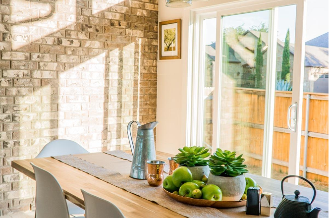 Sunlit dining table with green apples, succulents, and metal pitcher near brick wall and sliding glass door.