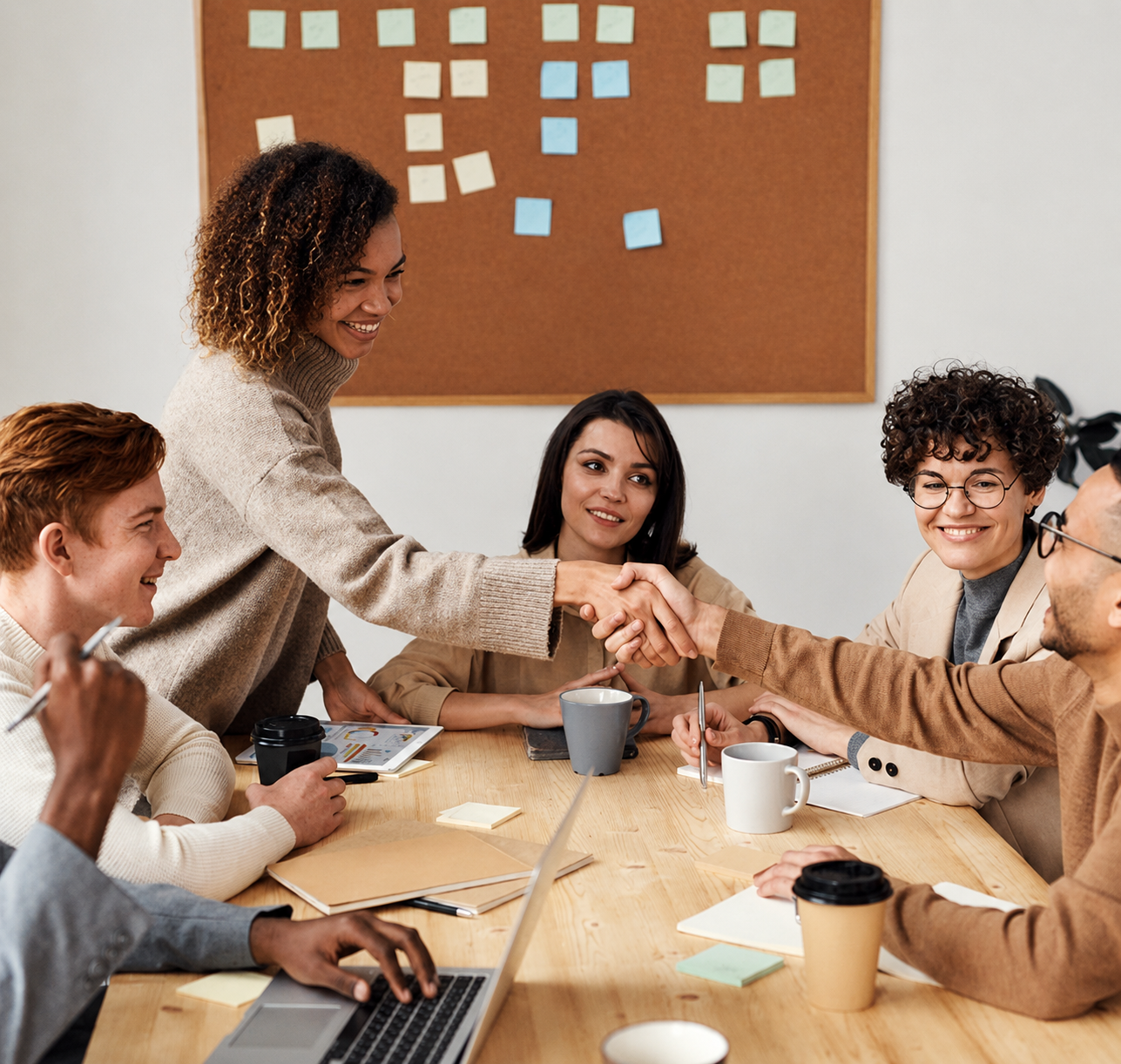 Five diverse coworkers greet with a handshake in a bright room with laptops and coffee cups.