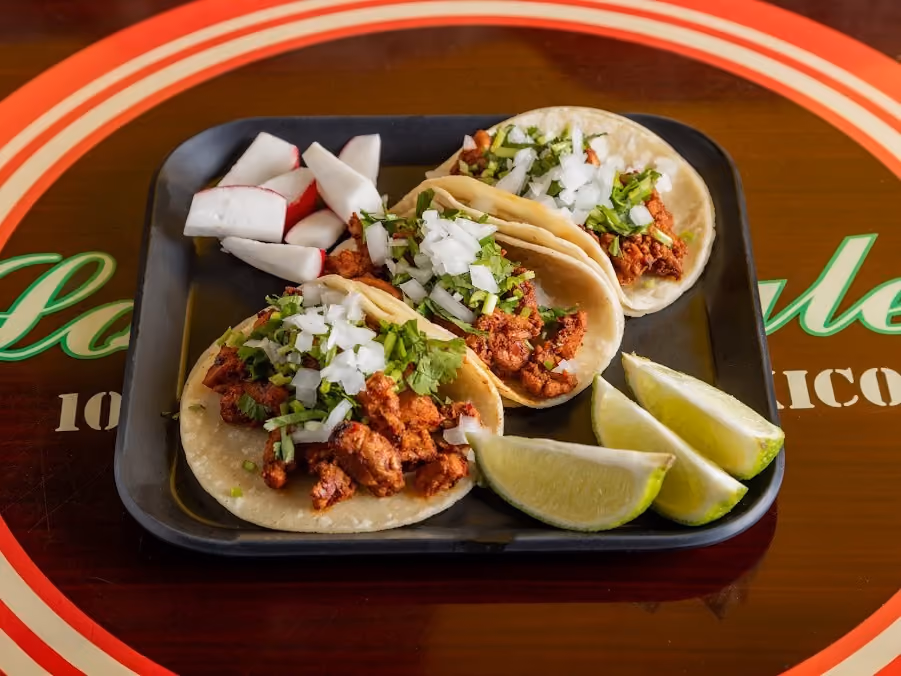 Three tacos with seasoned meat, chopped onion, and cilantro served with lime wedges and radish slices on a black tray.