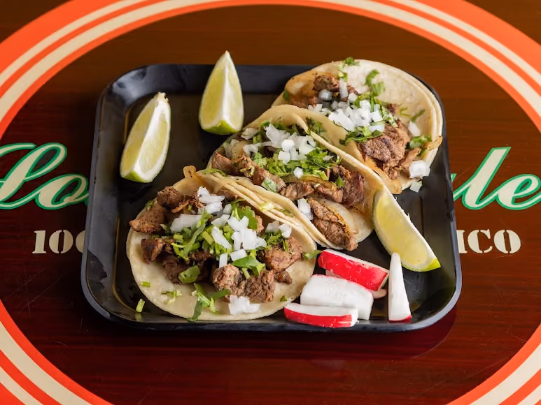 Three tacos with grilled meat, chopped onions, and cilantro served with lime wedges and radish slices on a black plate.