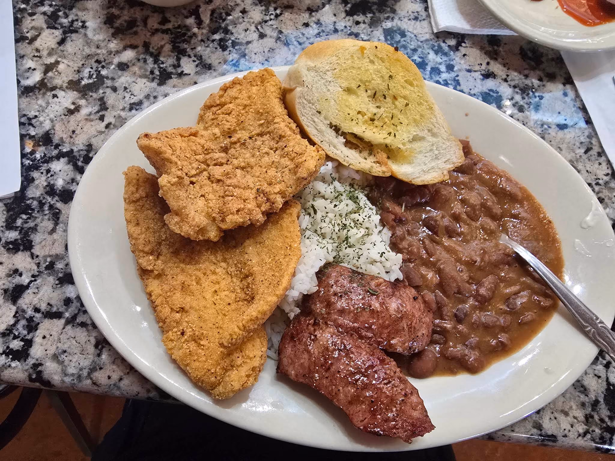 Plate with fried catfish, red beans, white rice, sausage link, and a slice of garlic bread.