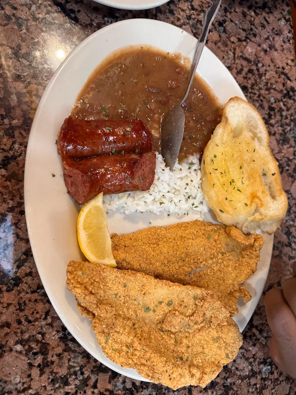 Plate with fried fish, sausage, dirty rice with gravy, garlic toast, and lemon wedge on a granite table.