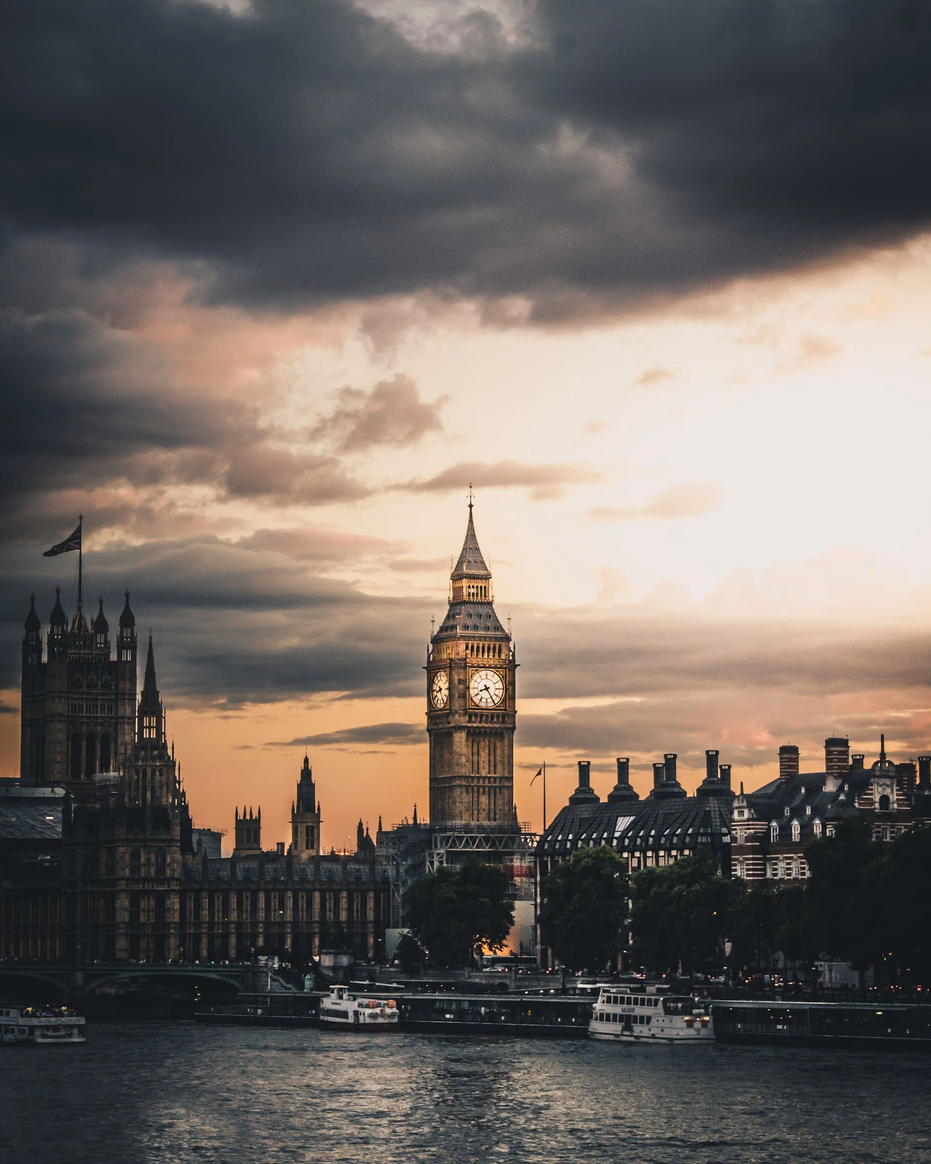 View of Big Ben and the Houses of Parliament in London at sunset with a partly cloudy sky and boats on the River Thames.