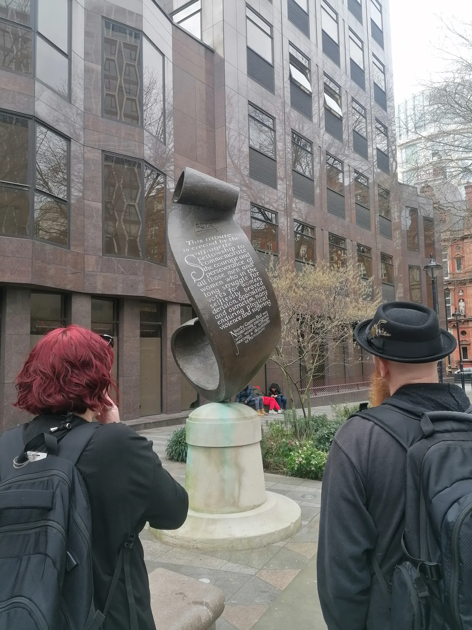 A public monument shaped like a scroll with an inscription commemorating the courage and perseverance of women in the suffragette movement, located outdoors in a city plaza, with two people viewing it from behind.