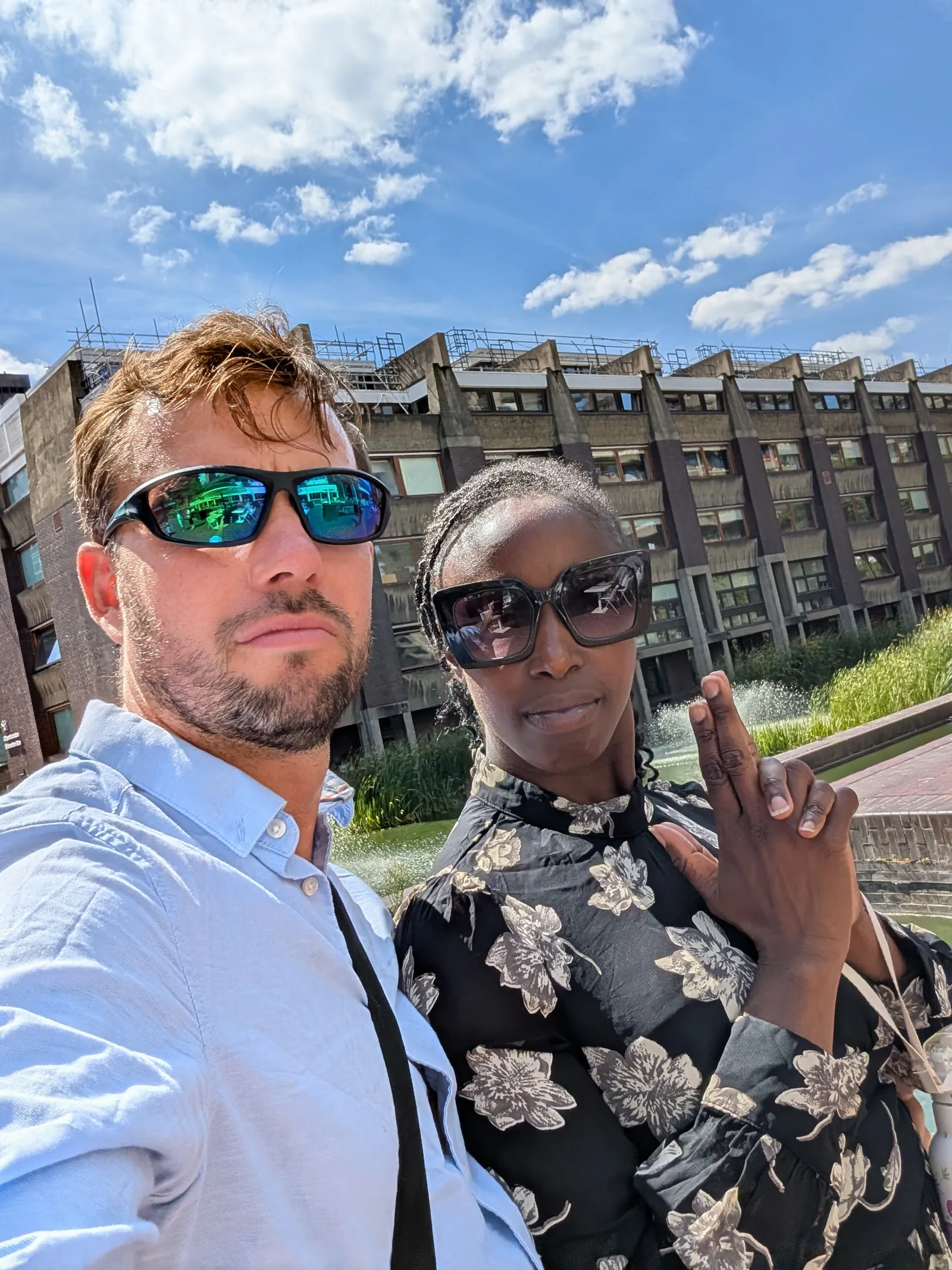 A man and woman outdoors in bright sunlight posing for a selfie in front of a modern building with water fountains and greenery. The man wears a light blue shirt and blue reflective sunglasses, and the woman wears a black floral patterned top and large black sunglasses, holding her hands together in a gesture.