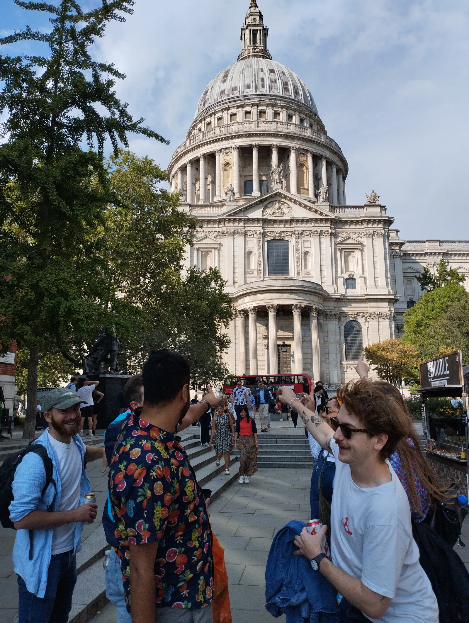 Group of young people outdoors laughing and pointing towards St. Paul's Cathedral in London on a clear day, with trees and a red double-decker bus in the background.