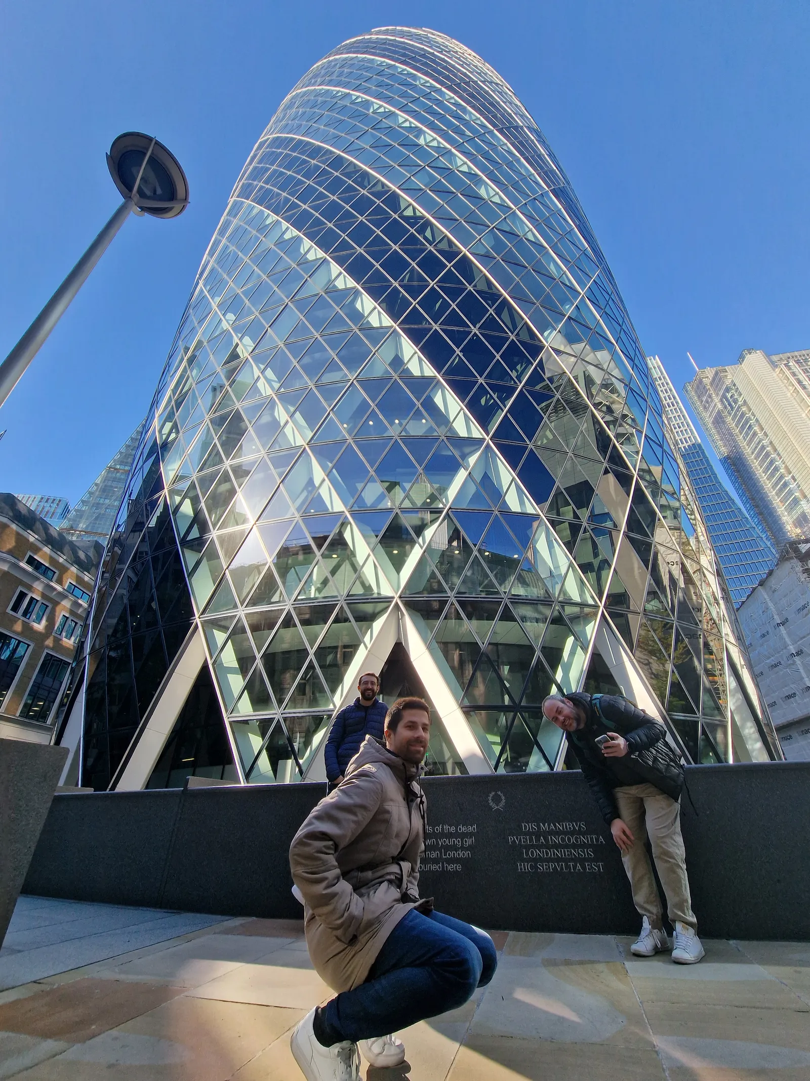 Three men posing in front of the Gherkin building in London on a clear day, with its distinctive glass and steel spiral pattern reflecting the sky and surrounding buildings.