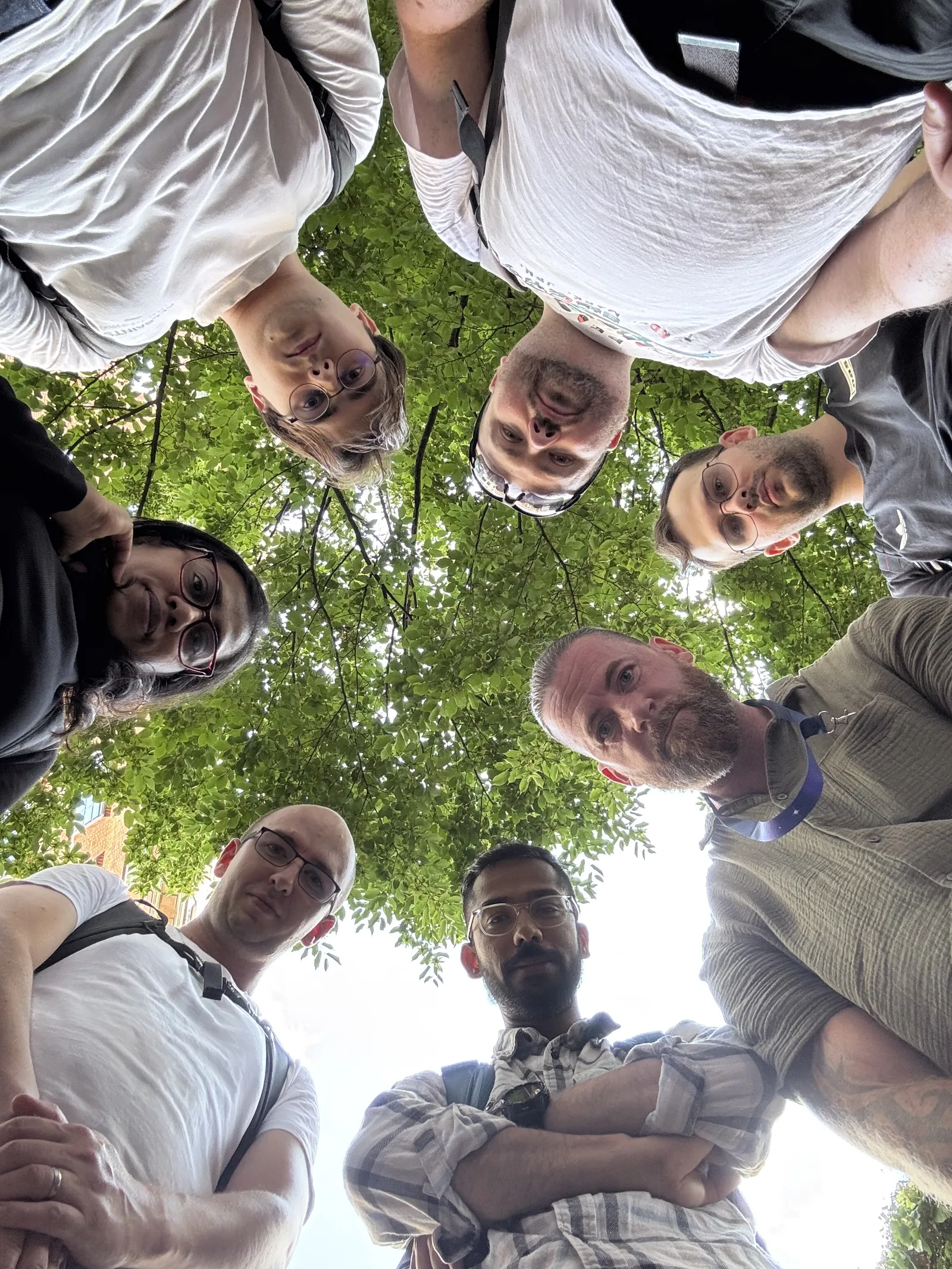 A group of seven adults standing in a circle outdoors looking down at the camera, with green leafy tree branches visible above them.