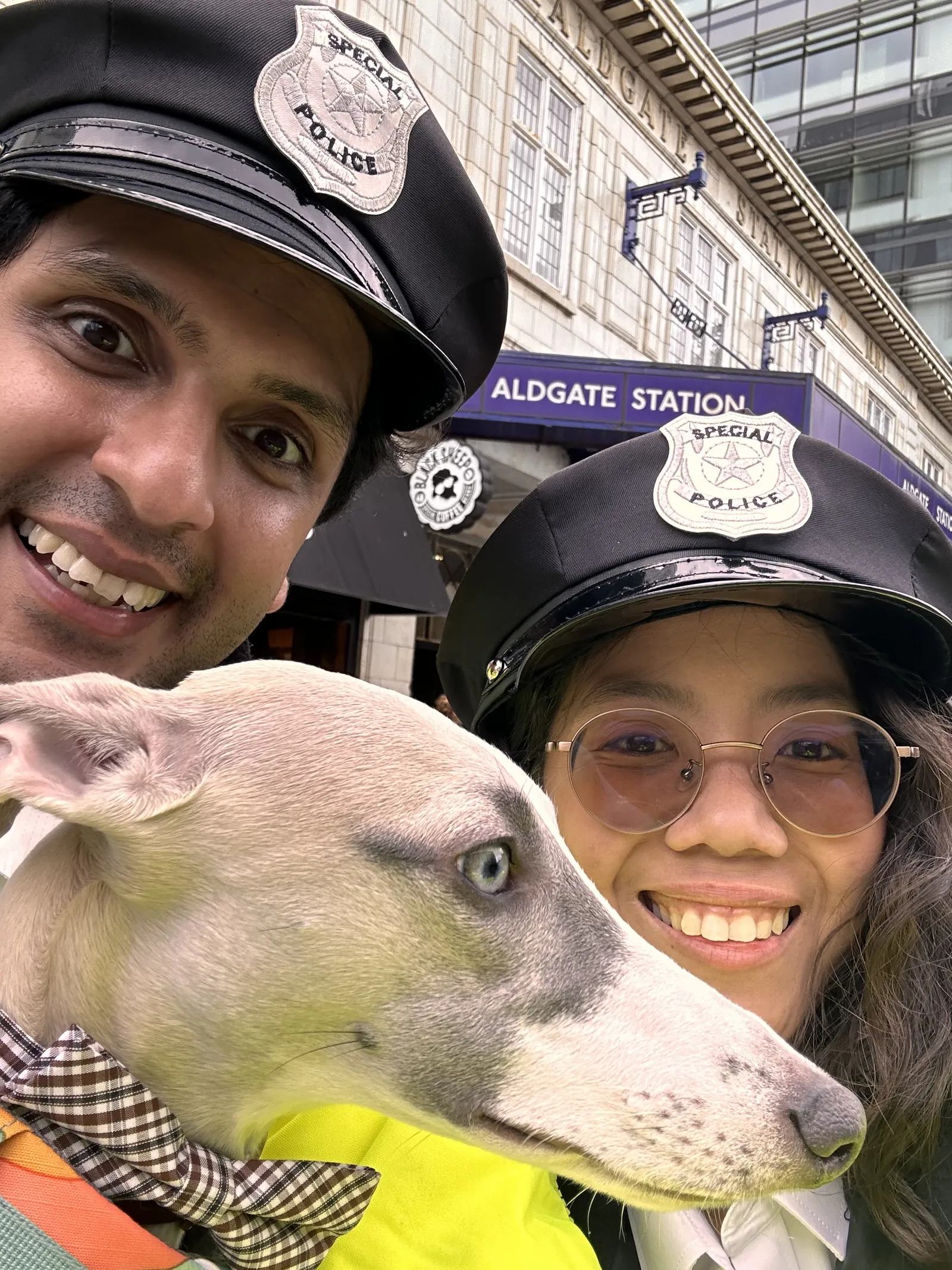 Two smiling people wearing black police hats with special police badges, posing closely with a grey and white dog wearing a checkered bow tie, in front of Aldgate Station.