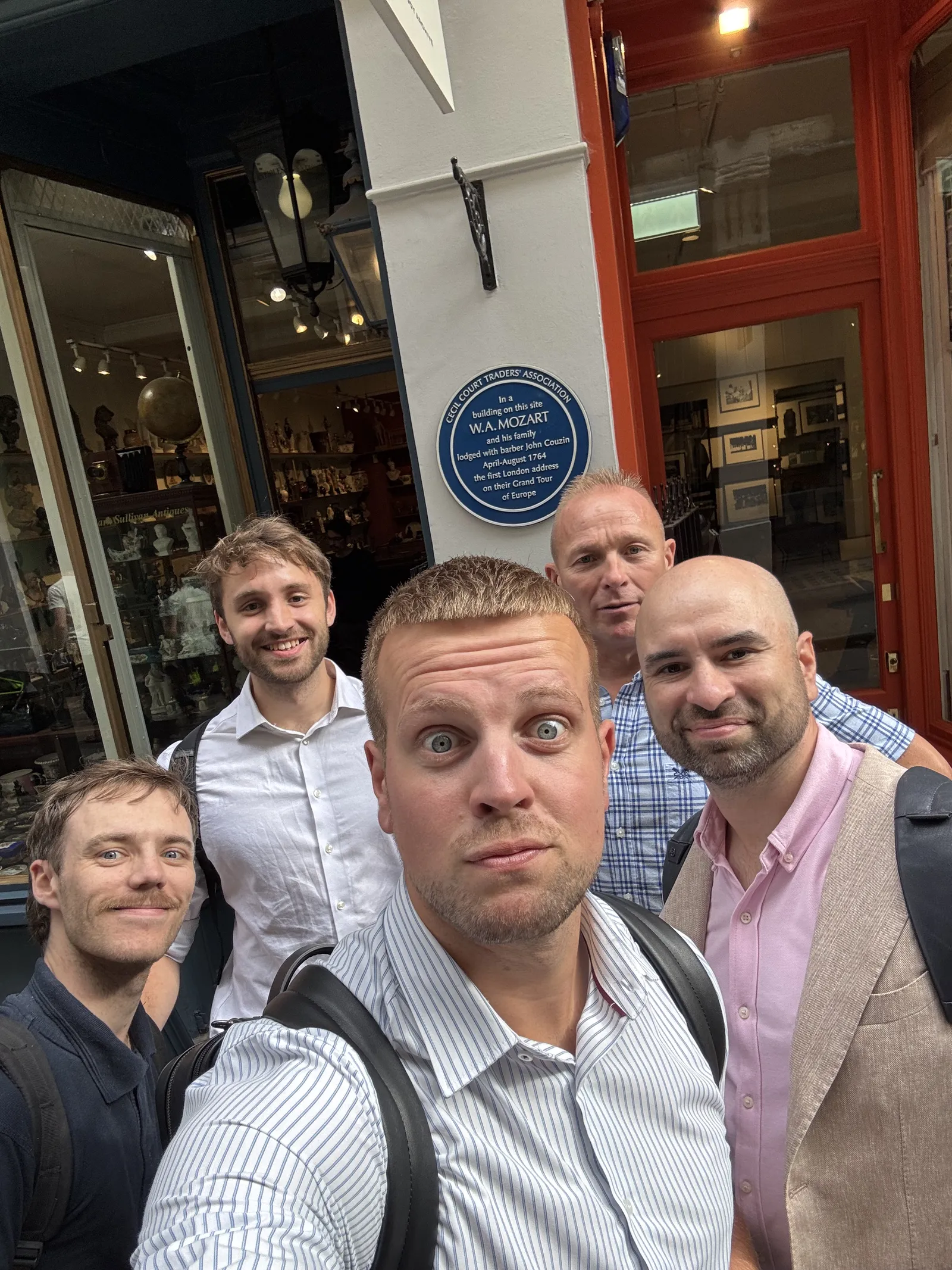 Five men taking a selfie outside a shop entrance with a blue plaque on the wall behind them noting that W. A. Mozart stayed there in 1764.